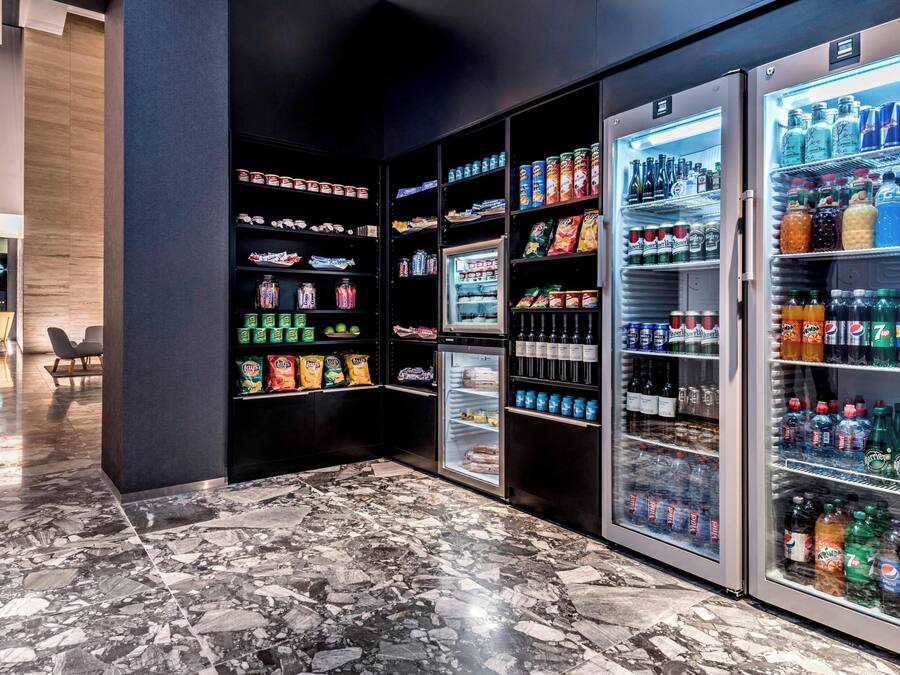 A modern hotel lobby with marble flooring, a seating area with chairs, and a row of beverage vending machines stocked with various drinks and snacks.