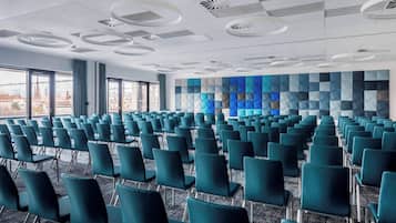 Une salle de conférence avec des rangées de chaises de couleur turquoise, un mur orné de motifs et de grandes fenêtres.