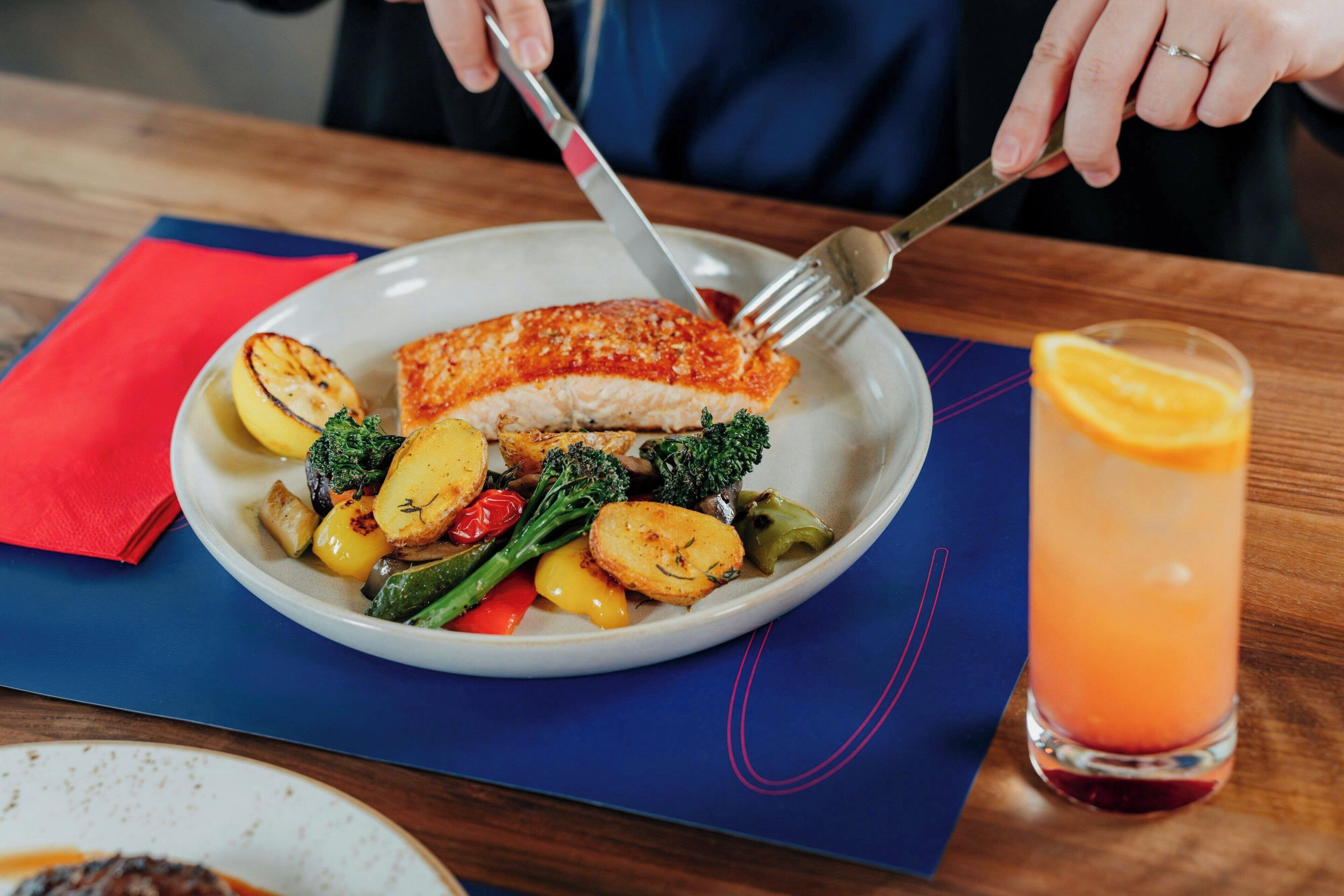 Un plato de comida con un trozo de pescado, verduras y un vaso de bebida sobre una mesa.