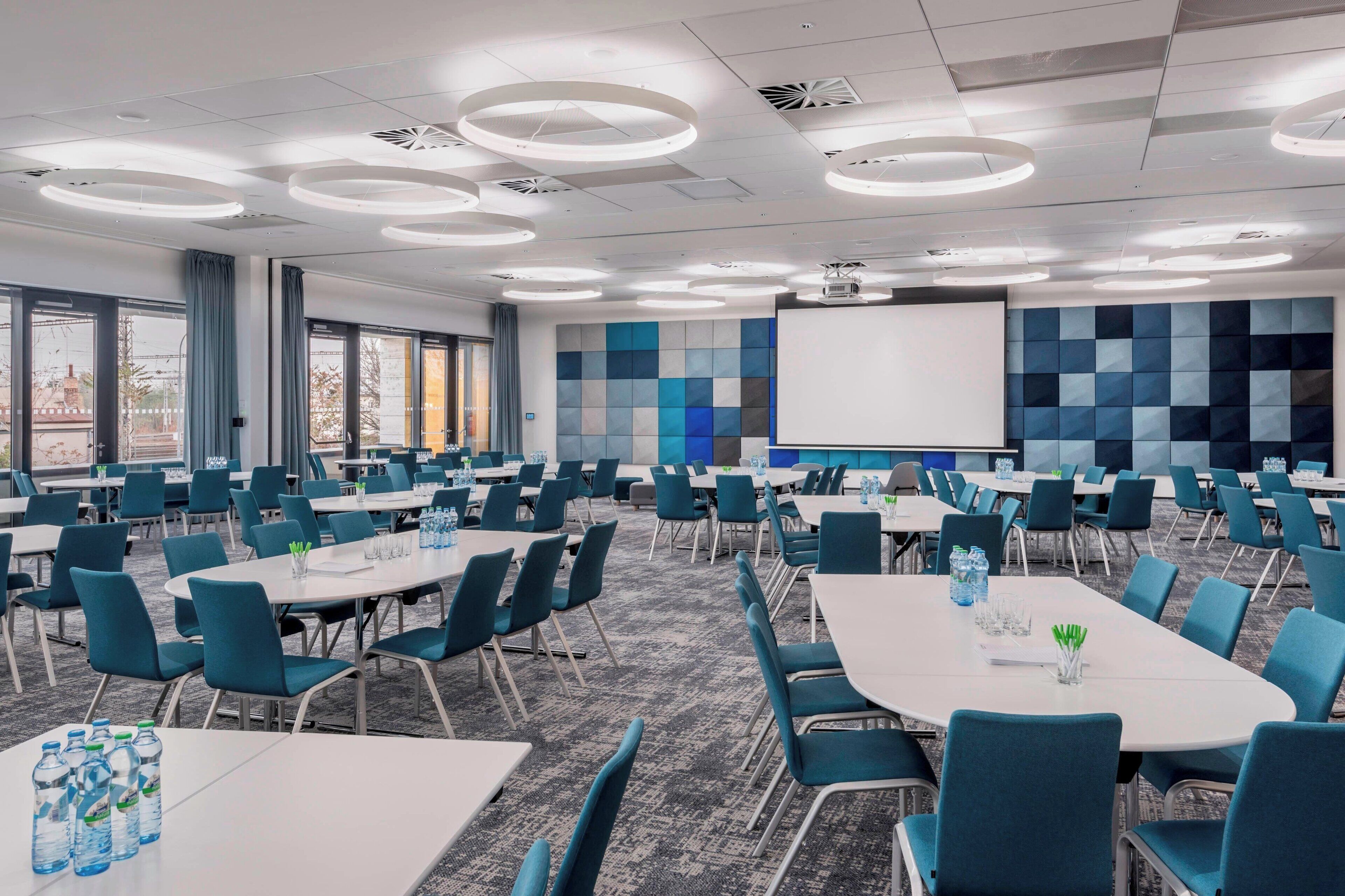 A conference room with multiple tables and chairs, a projector screen, and a large window with curtains.