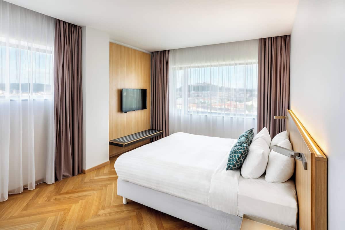 Une chambre d’hôtel avec un grand lit, un téléviseur fix é au mur et une vue sur la ville par les fenêtres.