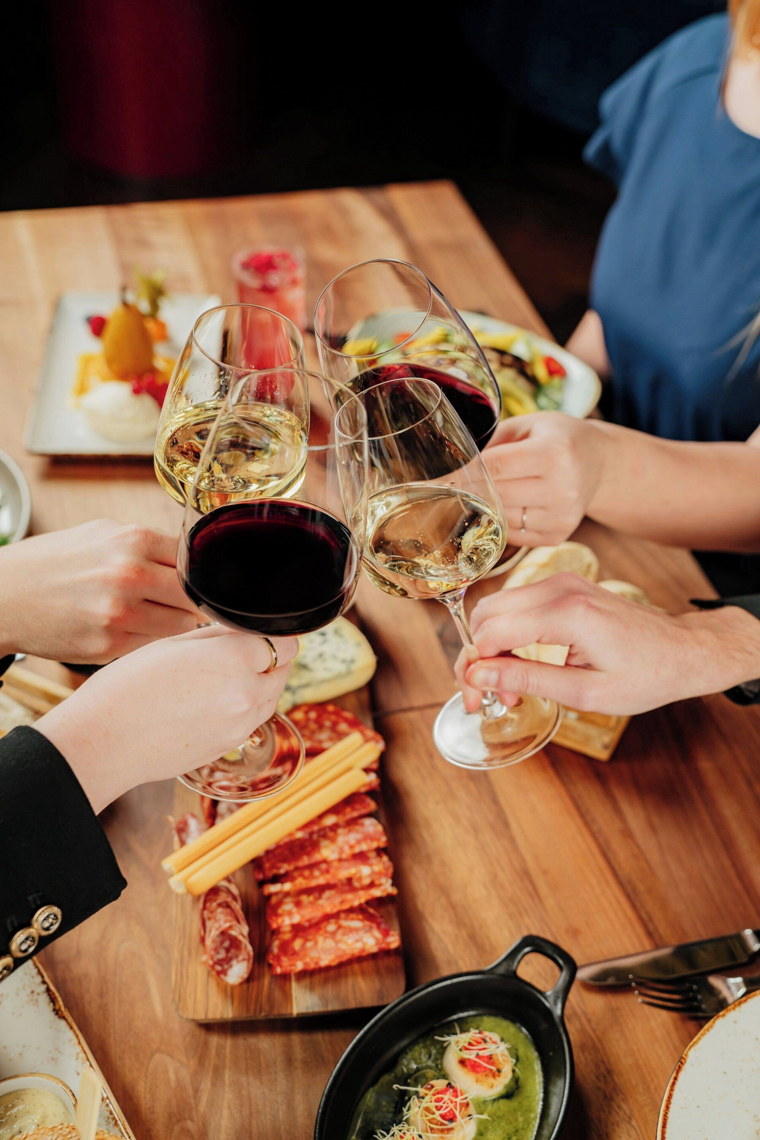 A group of people toasting with wine glasses at a wooden table with food and drinks.
