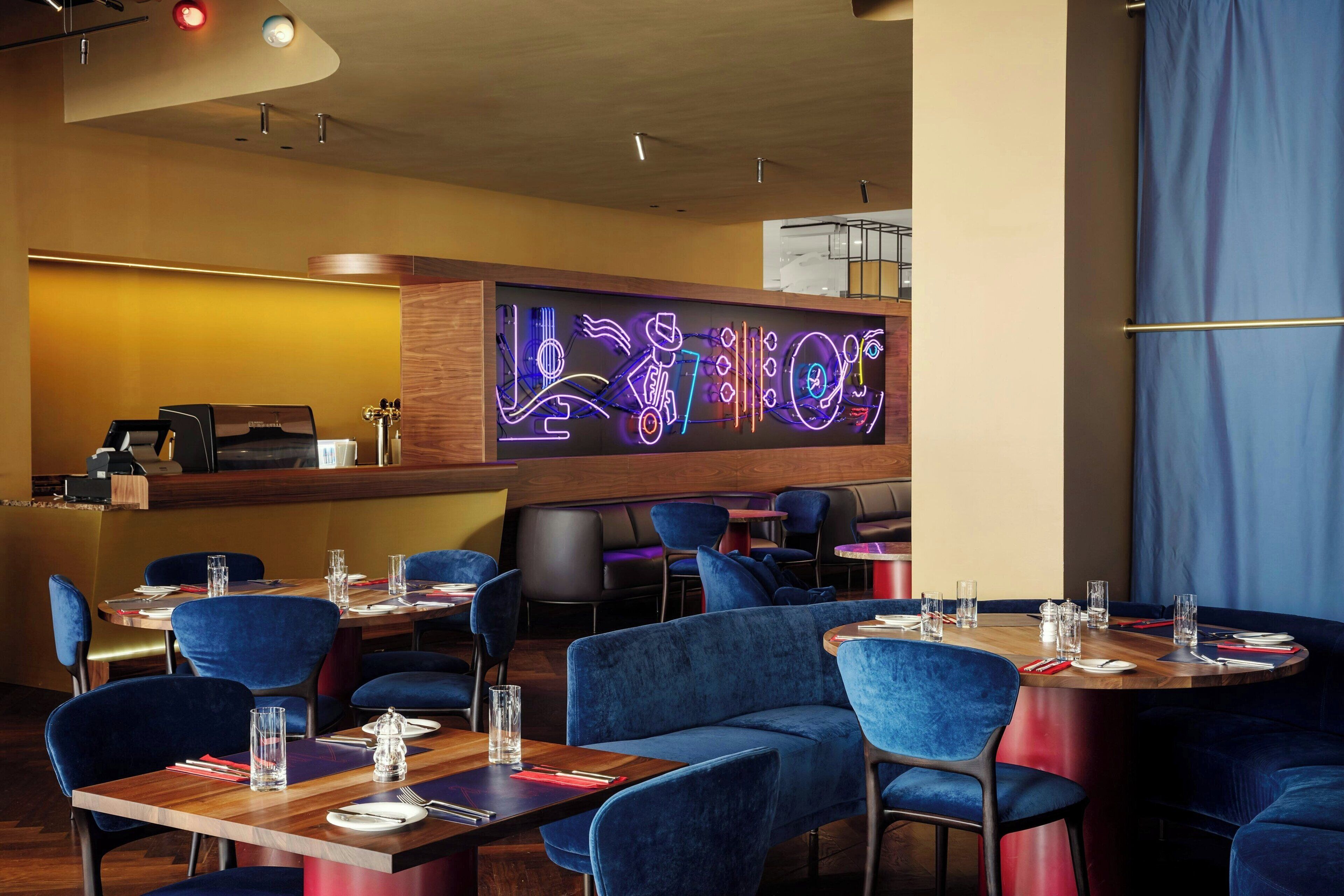 Un restaurant avec des chaises en velours bleu, des tables en bois et un panneau lumineux au néon sur le mur.