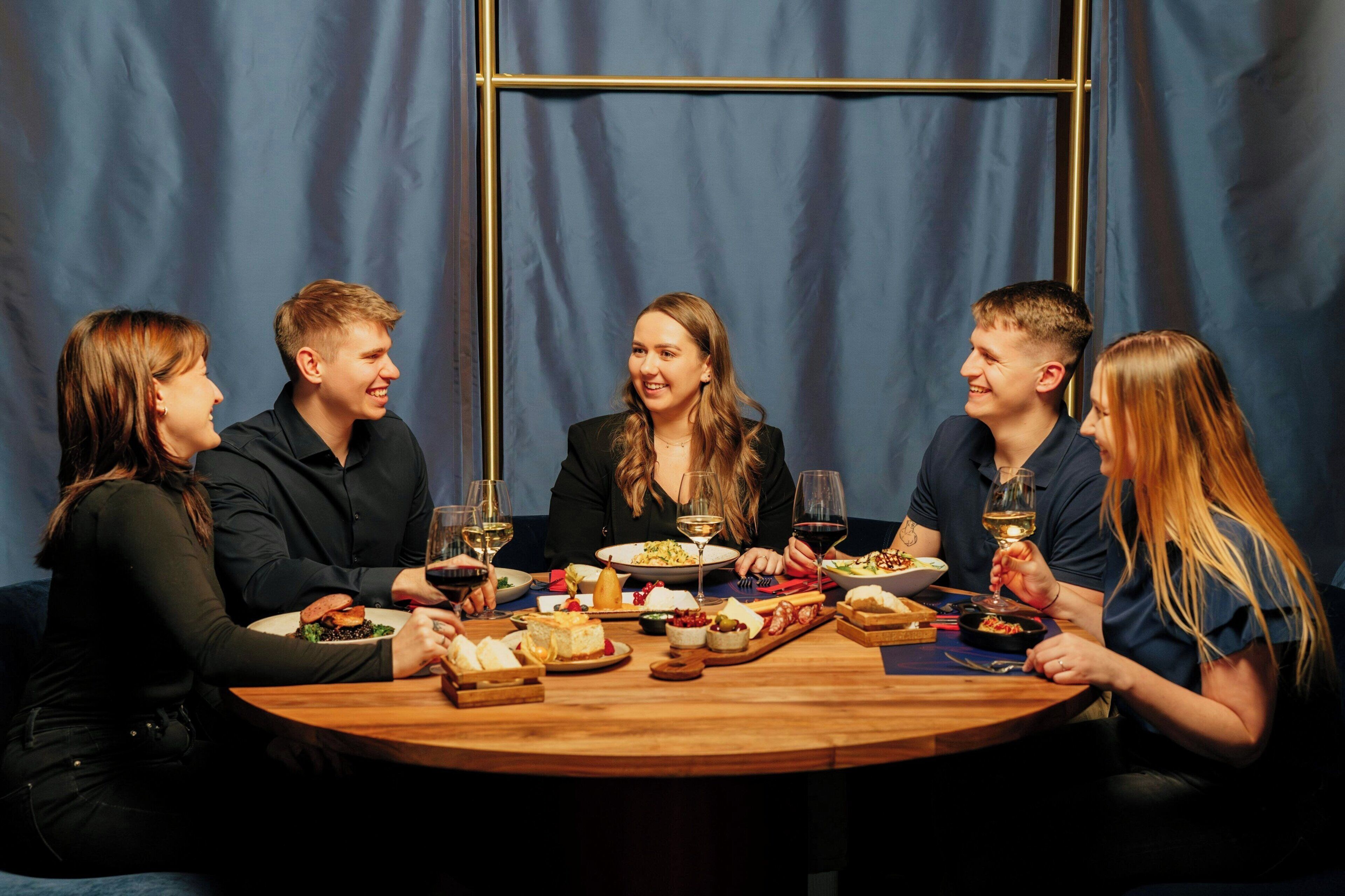 Un grupo de personas sentadas alrededor de una mesa de madera con varios platos y copas de vino, en un espacio con un telón azul de fondo.