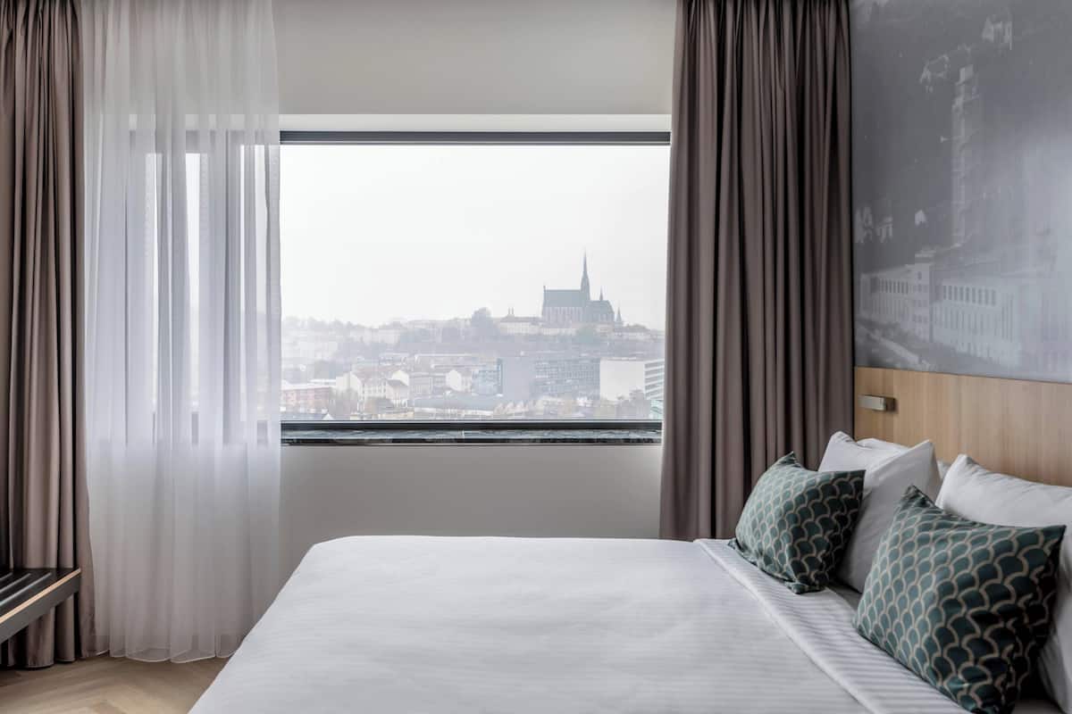 Une chambre d’hôtel avec un grand lit, une vue sur la ville par la fenêtre et un tableau représentant un paysage urbain accroché au mur.