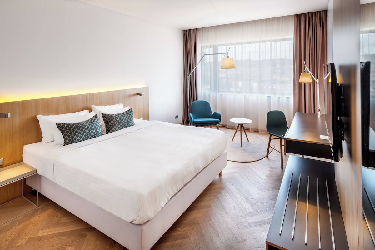 Une chambre d’hôtel avec un grand lit, un bureau avec une chaise, une télévision et une fenêtre avec des rideaux.