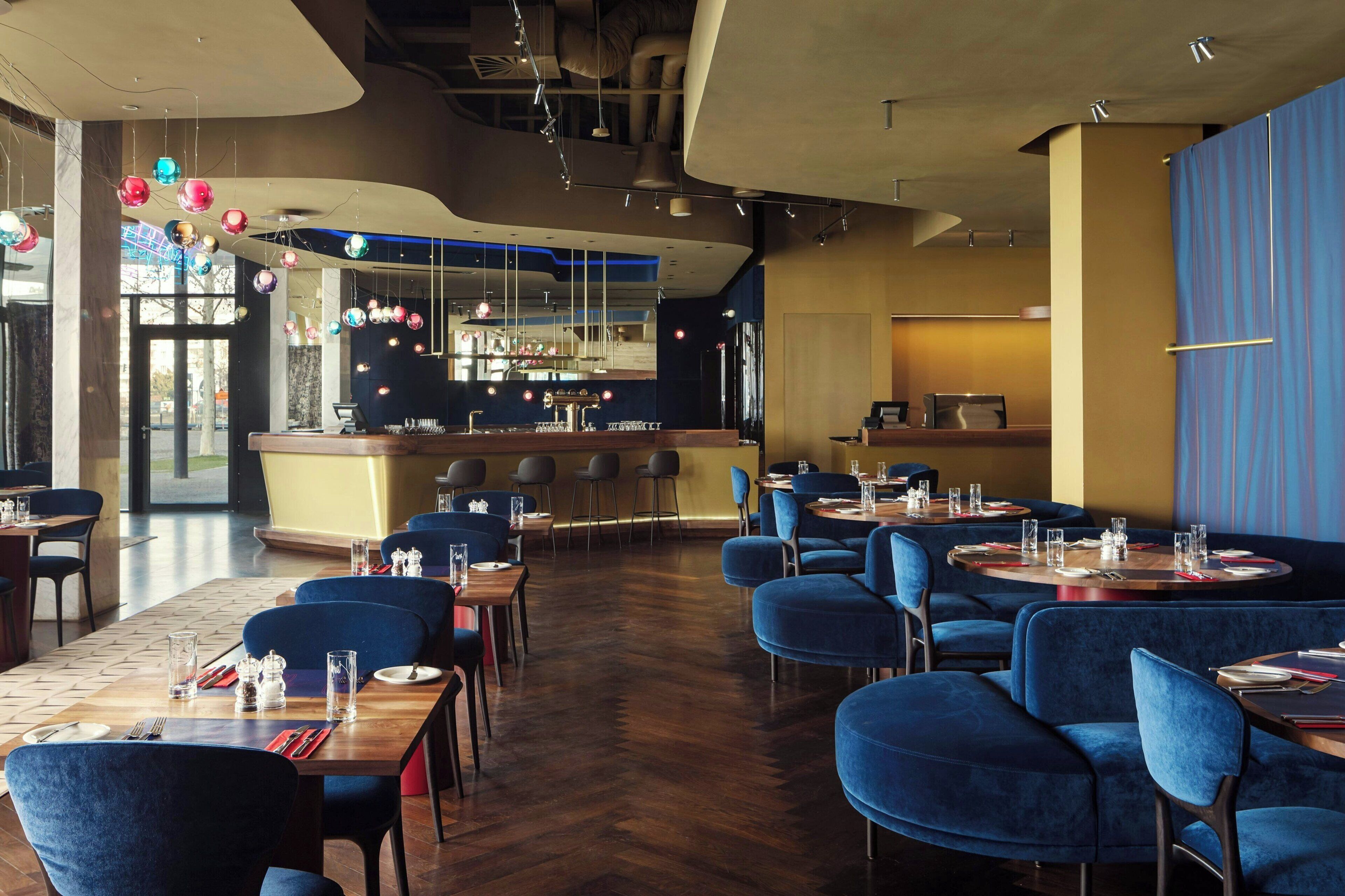 Ein Restaurant mit blauen Polstersesseln, Holztischen und hängenden, kugelförmigen Leuchten.