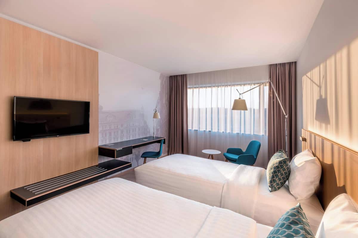 Une chambre d’hôtel avec deux lits, un bureau, une télévision, une lampe et une vue sur la ville par la fenêtre.