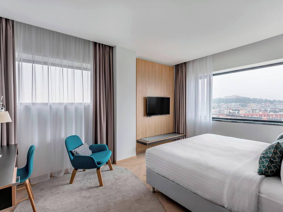 Habitación de hotel con cama, un escritorio con silla, televisor montado en la pared y un gran ventanal con vista a la ciudad.