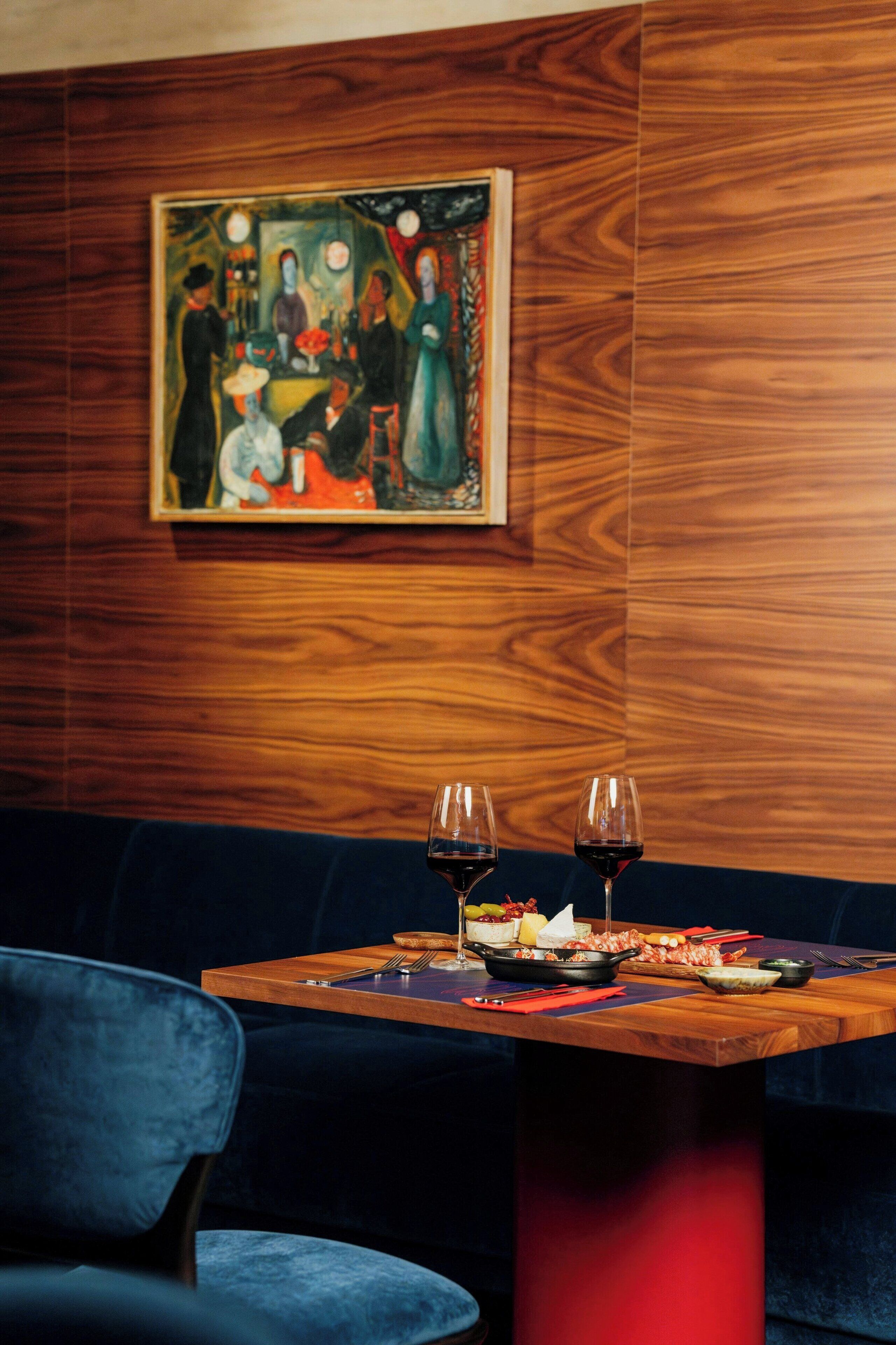 Uma sala de jantar com uma mesa de madeira posta para dois, um banco estofado azul e um quadro na parede.