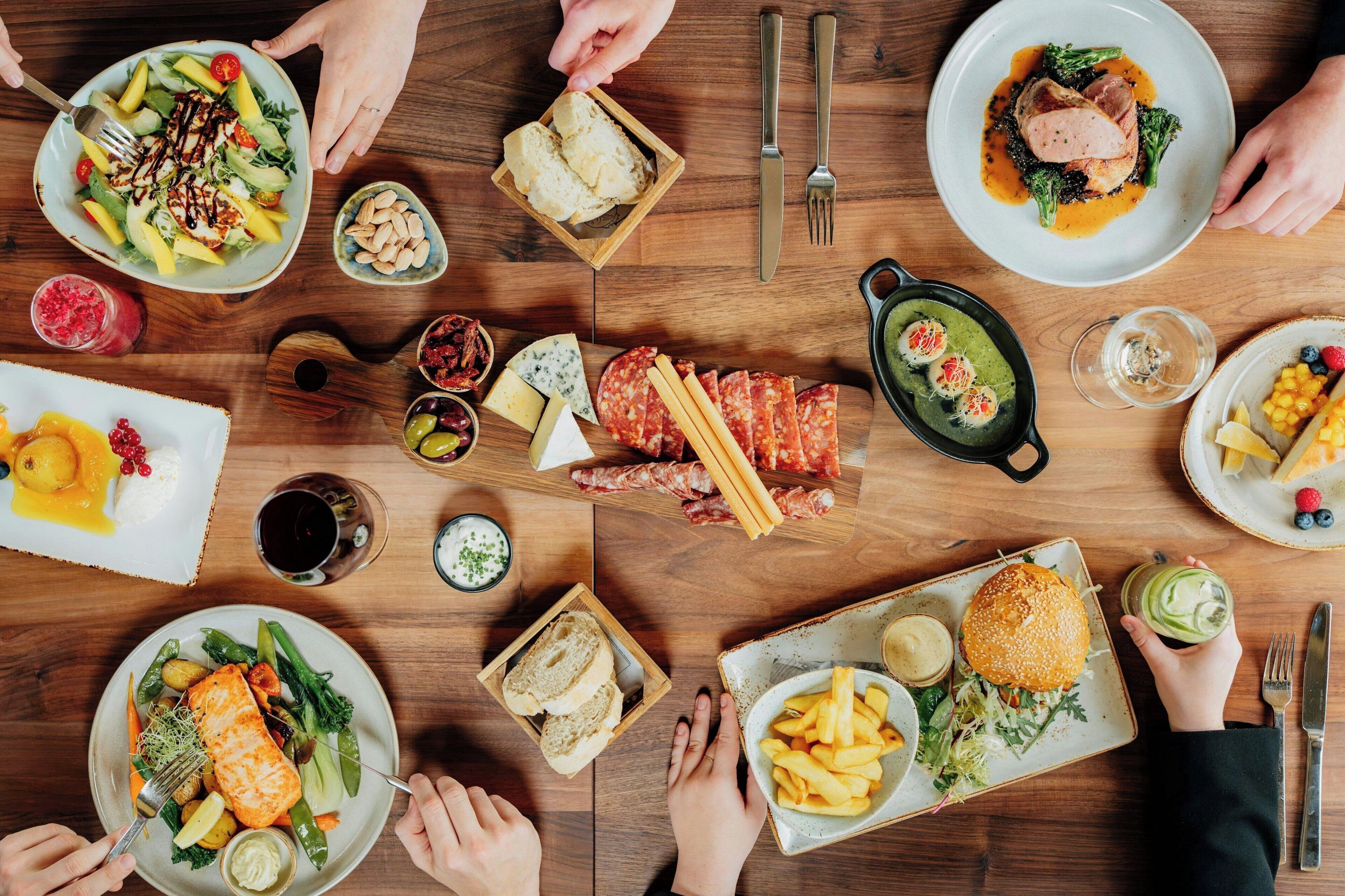 A dining table with various dishes, including salmon, steak, and vegetables, surrounded by people.