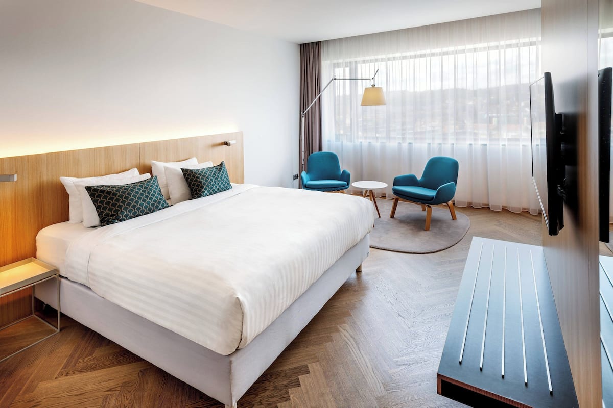 Une chambre d’hôtel moderne avec un grand lit, deux fauteuils bleus, une petite table ronde, un téléviseur et une grande fenêtre avec des rideaux légers.