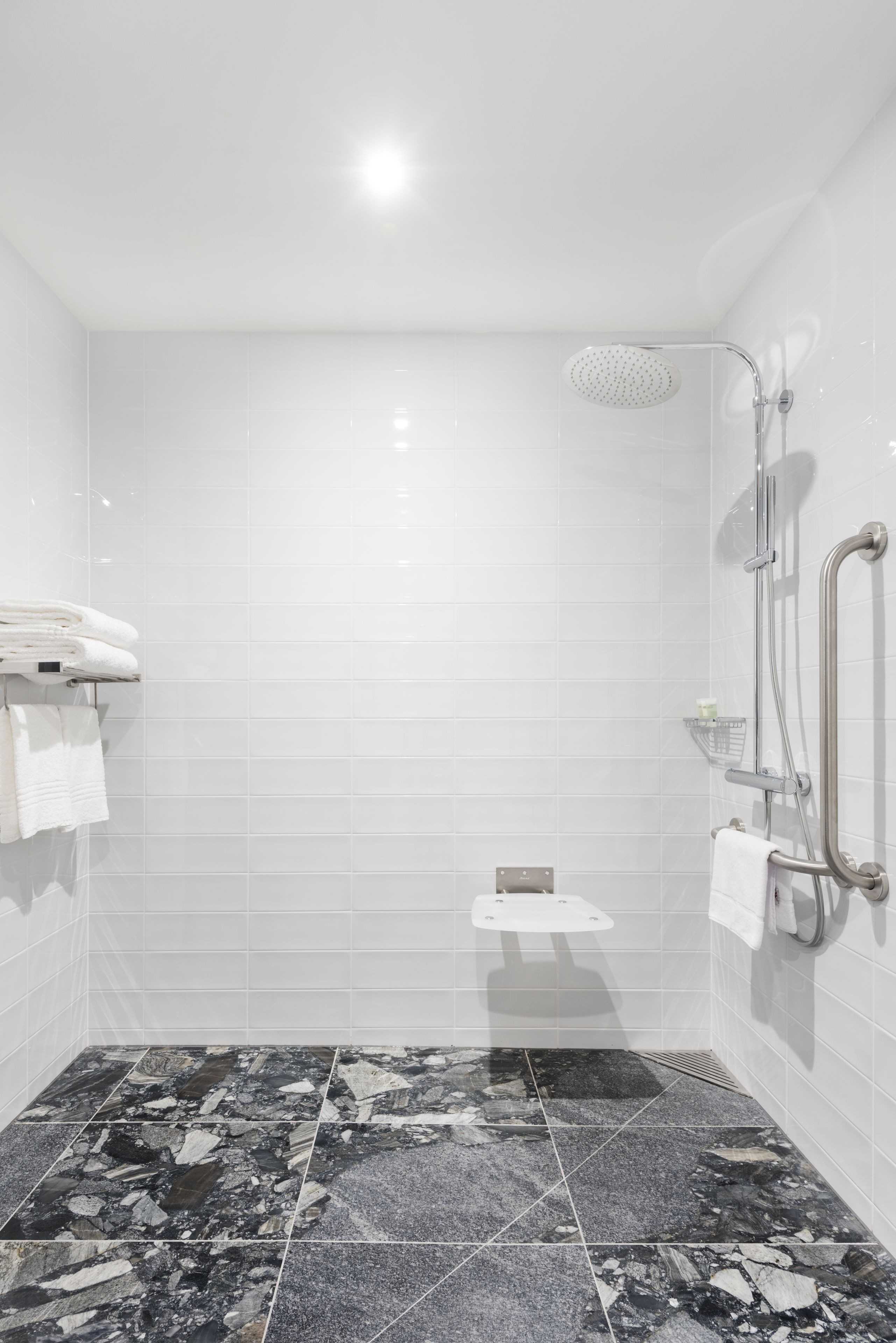 A modern bathroom with white tiled walls, a shower with a handheld showerhead, and a marble floor.