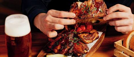 Una persona sosteniendo un gran trozo de carne, posiblemente costillas, con una porción de papas fritas y una cerveza en una mesa de madera.