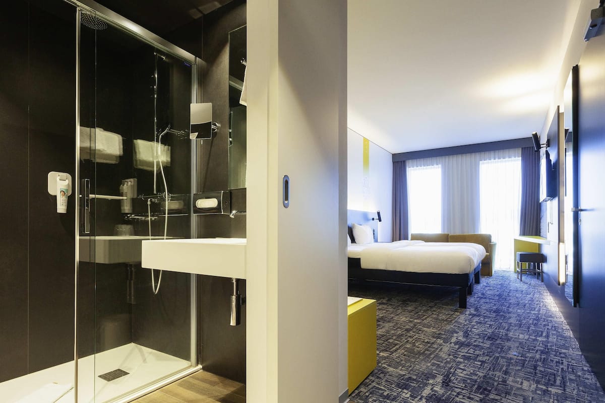 A hotel room with a glass-enclosed shower, a bed with a headboard, a bedside table, and a small stool.