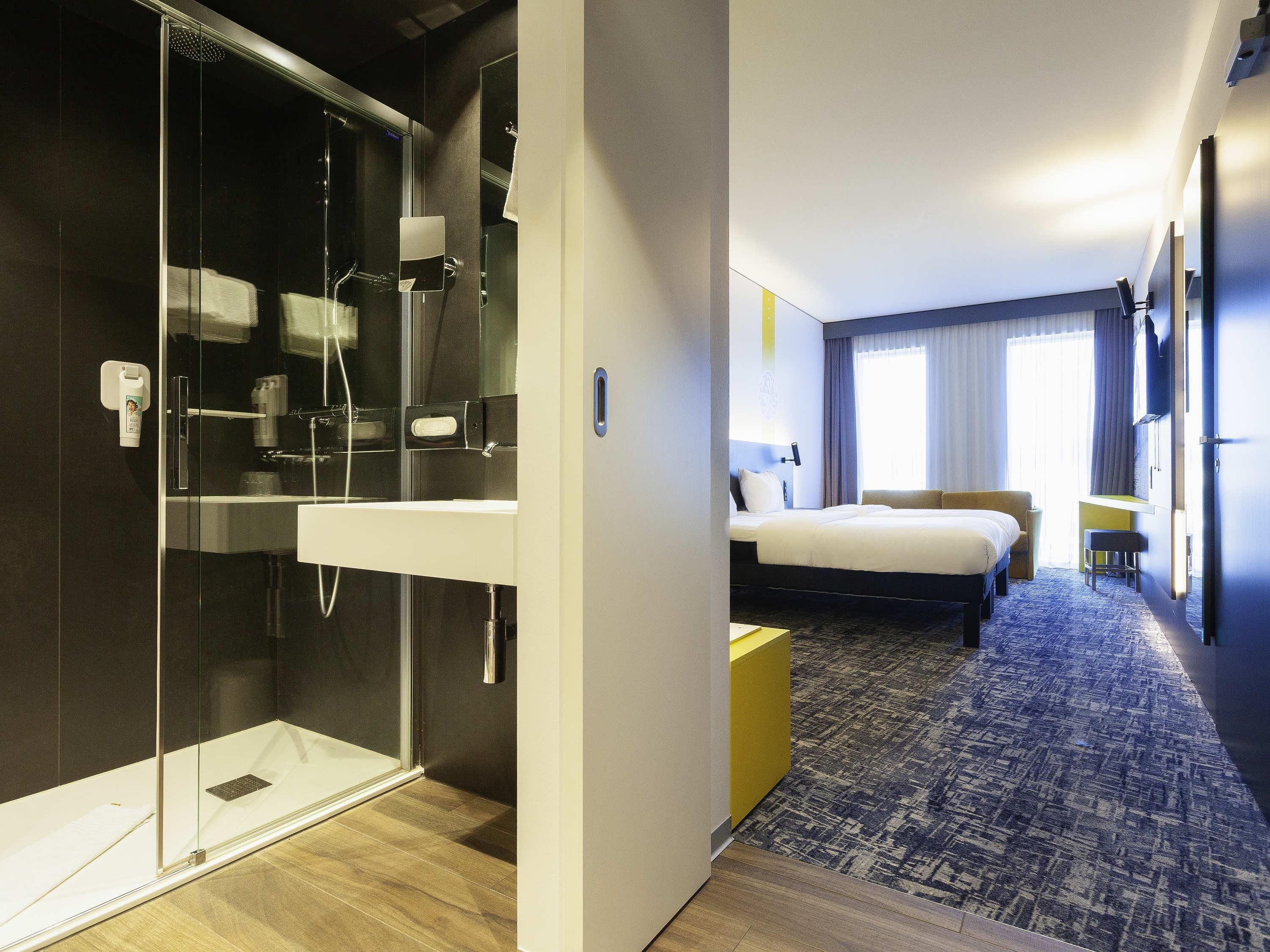 Une chambre d’hôtel dotée d’une douche en verre, d’un lit avec une tête de lit, d’une table de chevet et d’un petit tabouret.