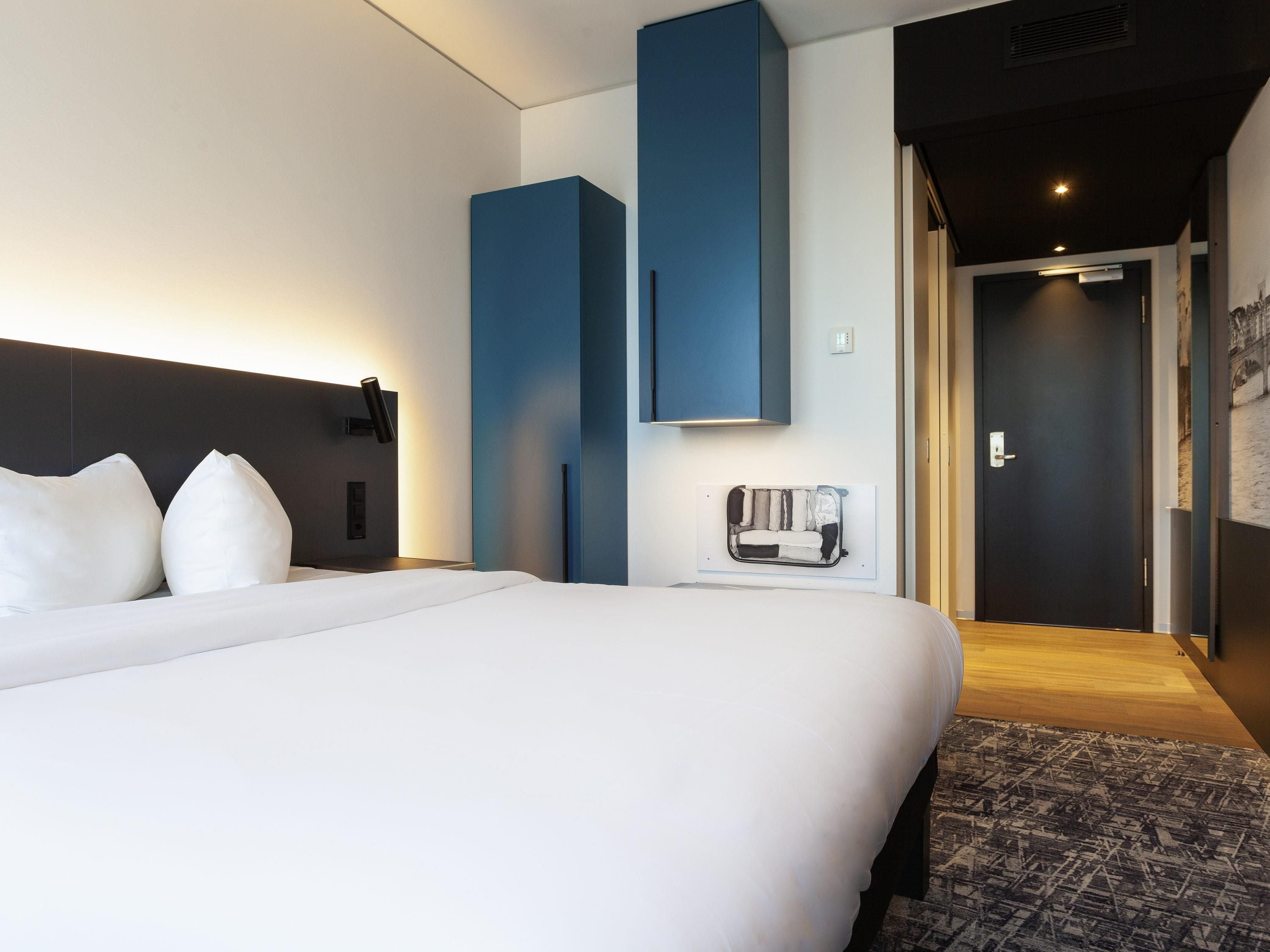 A modern hotel room with a large bed, a bedside table, a built-in bench with storage, a wall-mounted TV, and a door leading to another room.