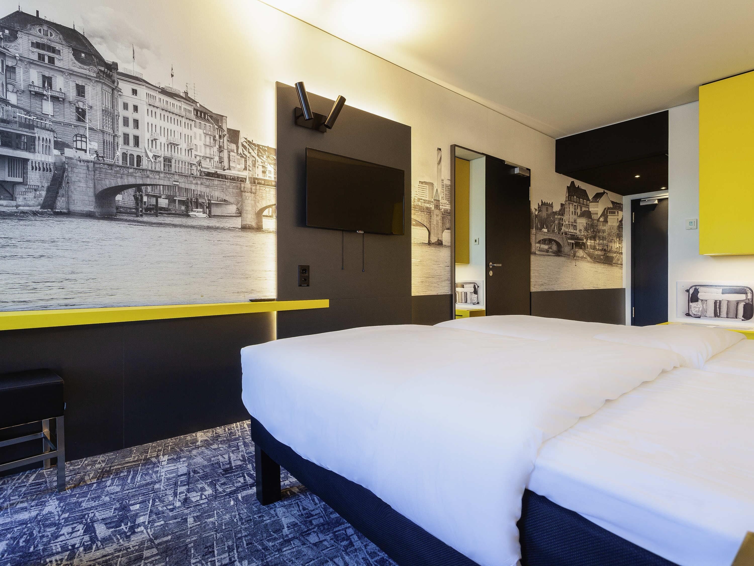 Ein Hotelzimmer mit einem Bett, einem an der Wand montierten Fernseher und einem großen Wandgemälde einer Stadtansicht.