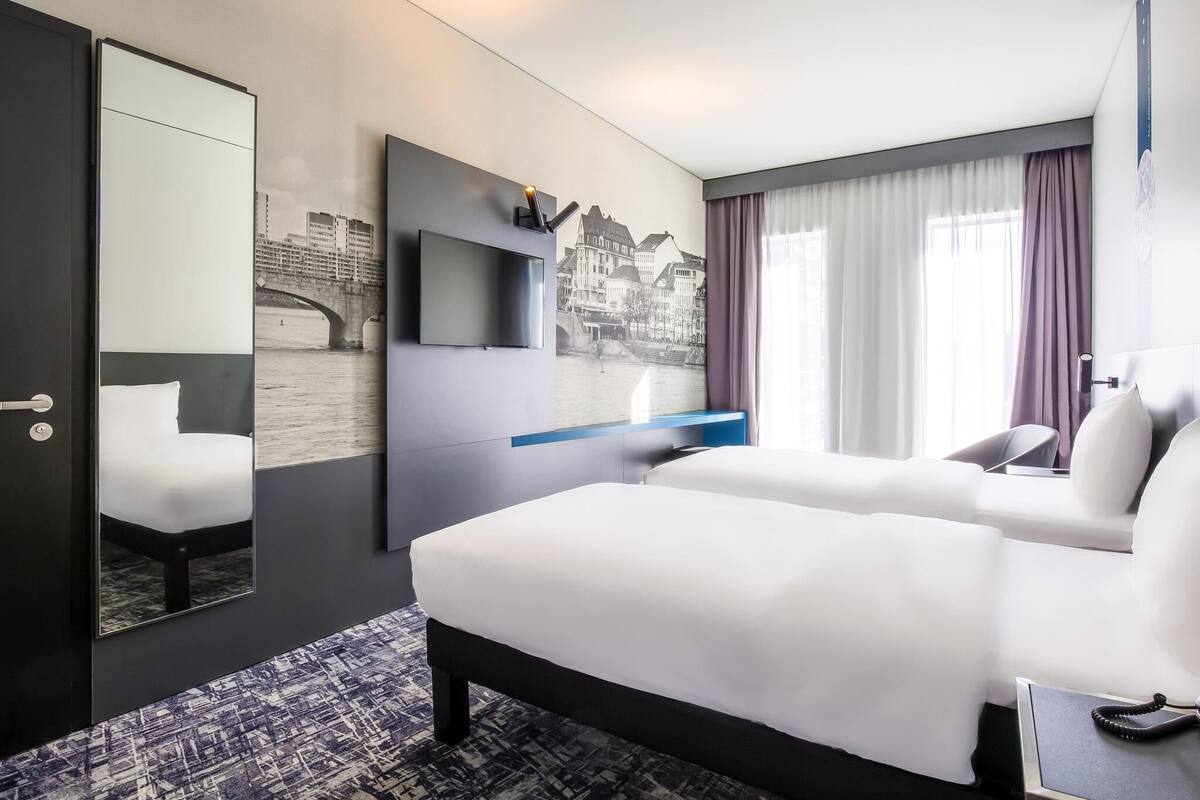 A hotel room with a bed, a desk, a chair, a mirror, and a window with curtains.