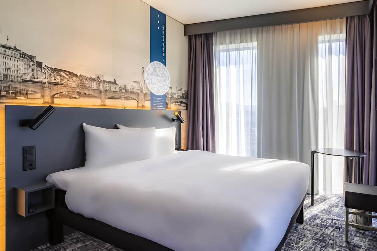 A hotel room with a large bed, a nightstand, a desk, a chair, and a view of a cityscape through the window.