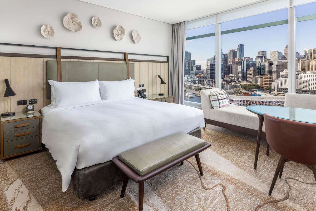 A modern hotel room with a large bed, a bench, a chair, and a view of a city skyline.