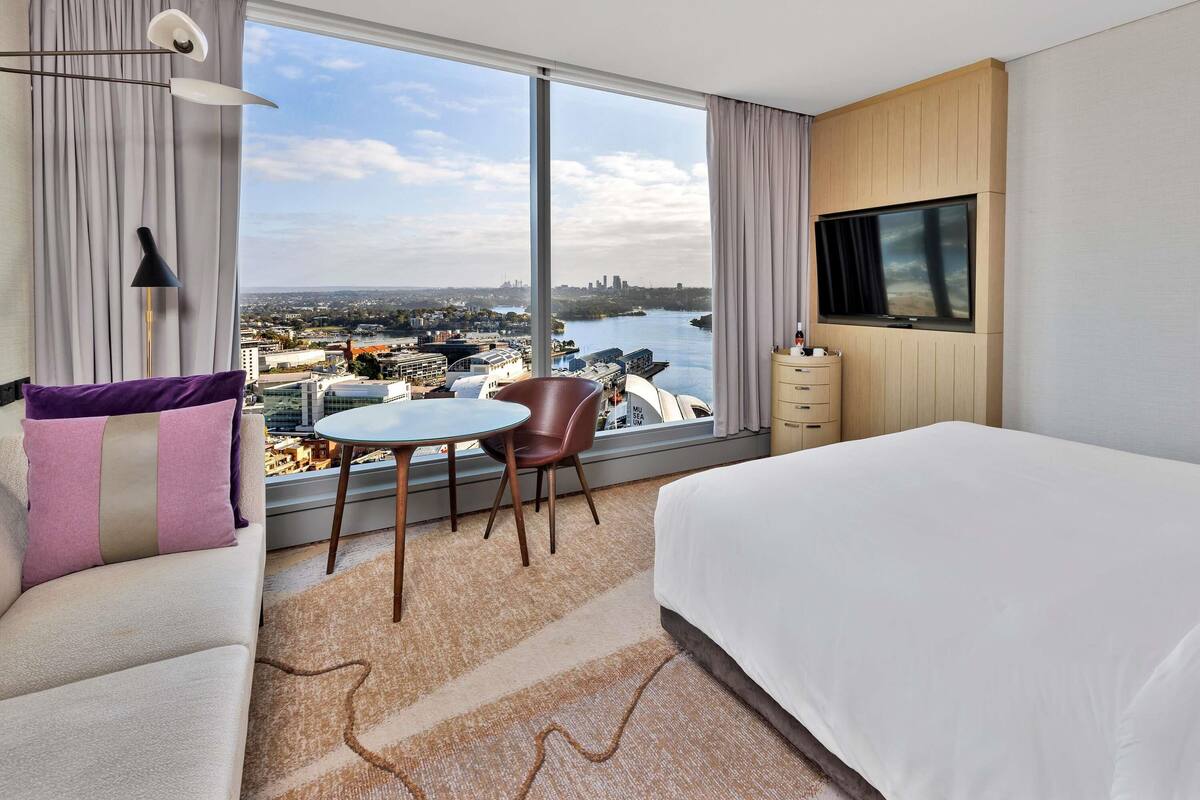 A hotel room with a large bed, a TV mounted on the wall, a dining table with chairs, and a view of a cityscape through large windows.