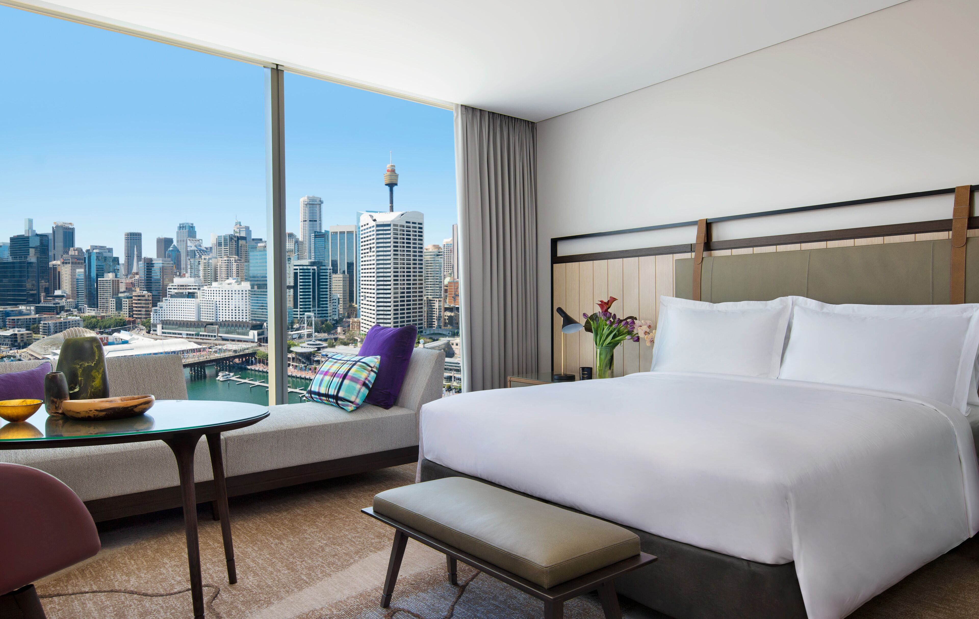 A modern hotel room with a large bed, a sofa, a small table, and a view of a city skyline.