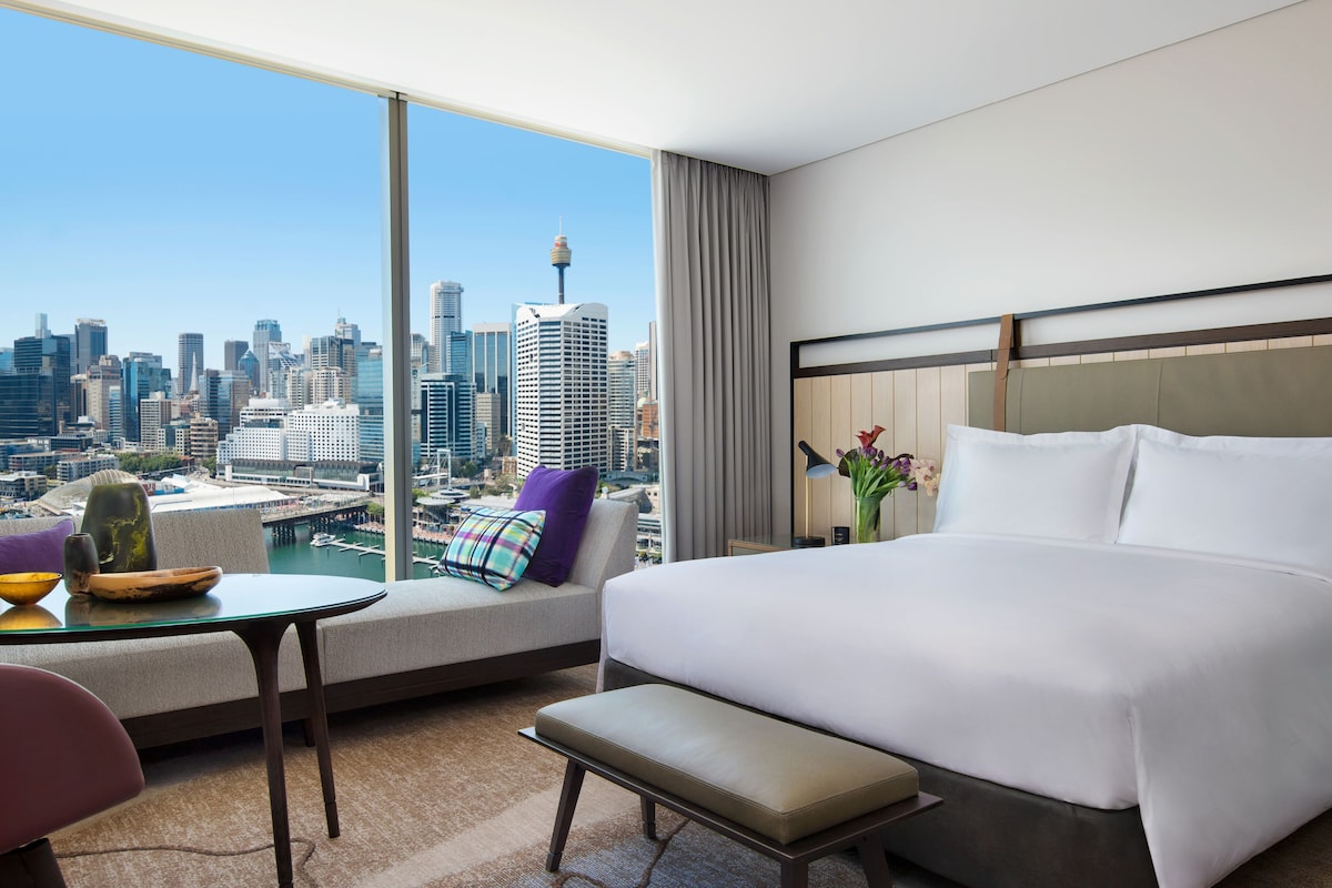 Habitación de hotel moderna con una cama grande, un sofá, una mesita y vistas a un horizonte urbano.