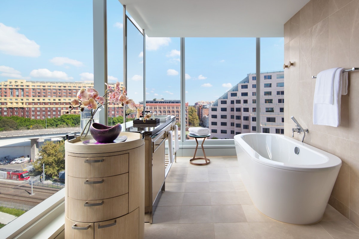 Un baño moderno con bañera, tocador con cajones, ventana con vistas a edificios y un pequeño taburete.
