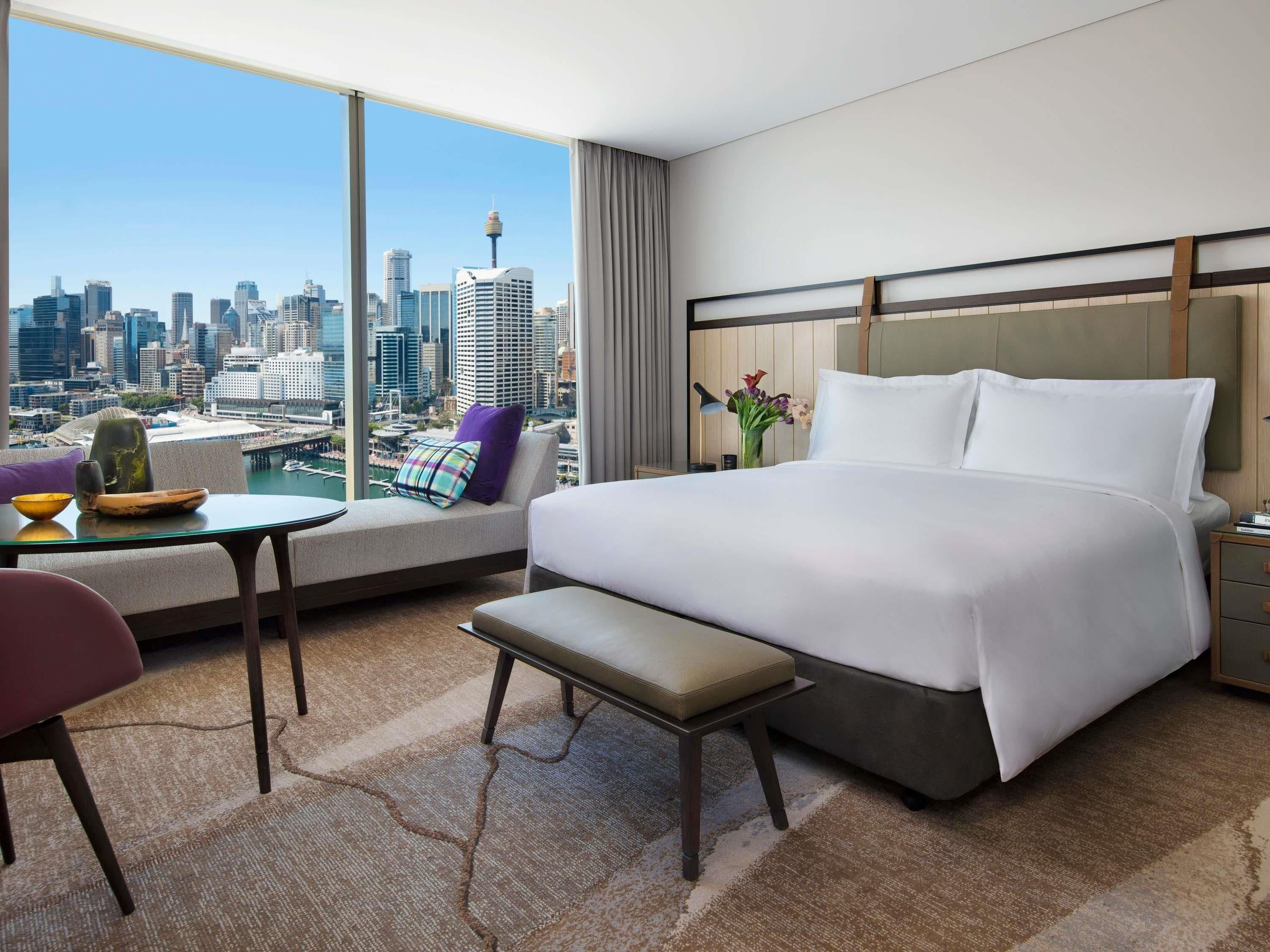 Habitación de hotel con una cama grande, una mesita, una silla y vistas a un horizonte urbano.