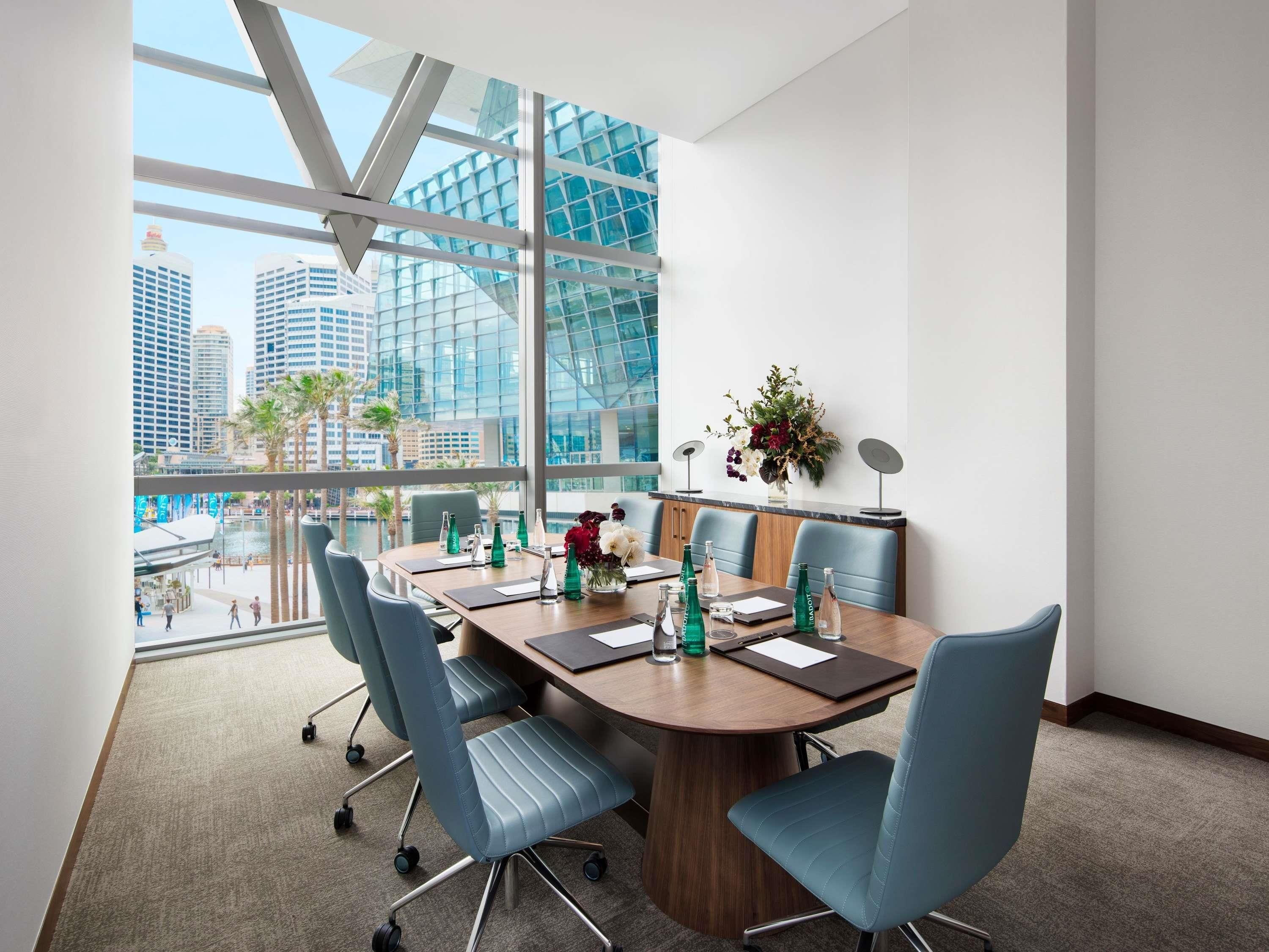 A modern conference room with a large table, blue chairs, and a view of city buildings through large windows.