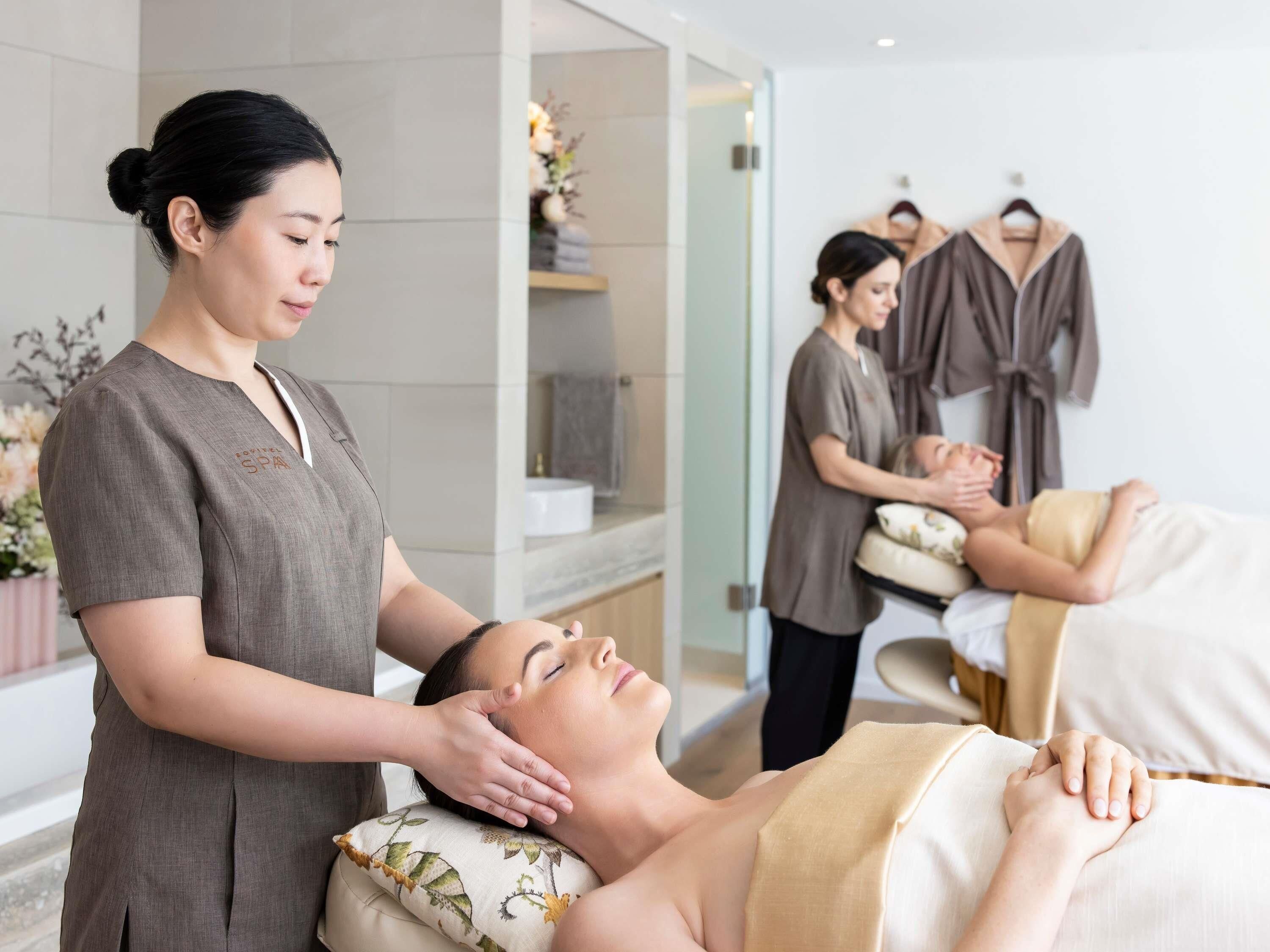 Un ambiente de spa con una camilla de masaje, una persona recibiendo un masaje y otra persona atendiendo a un cliente.