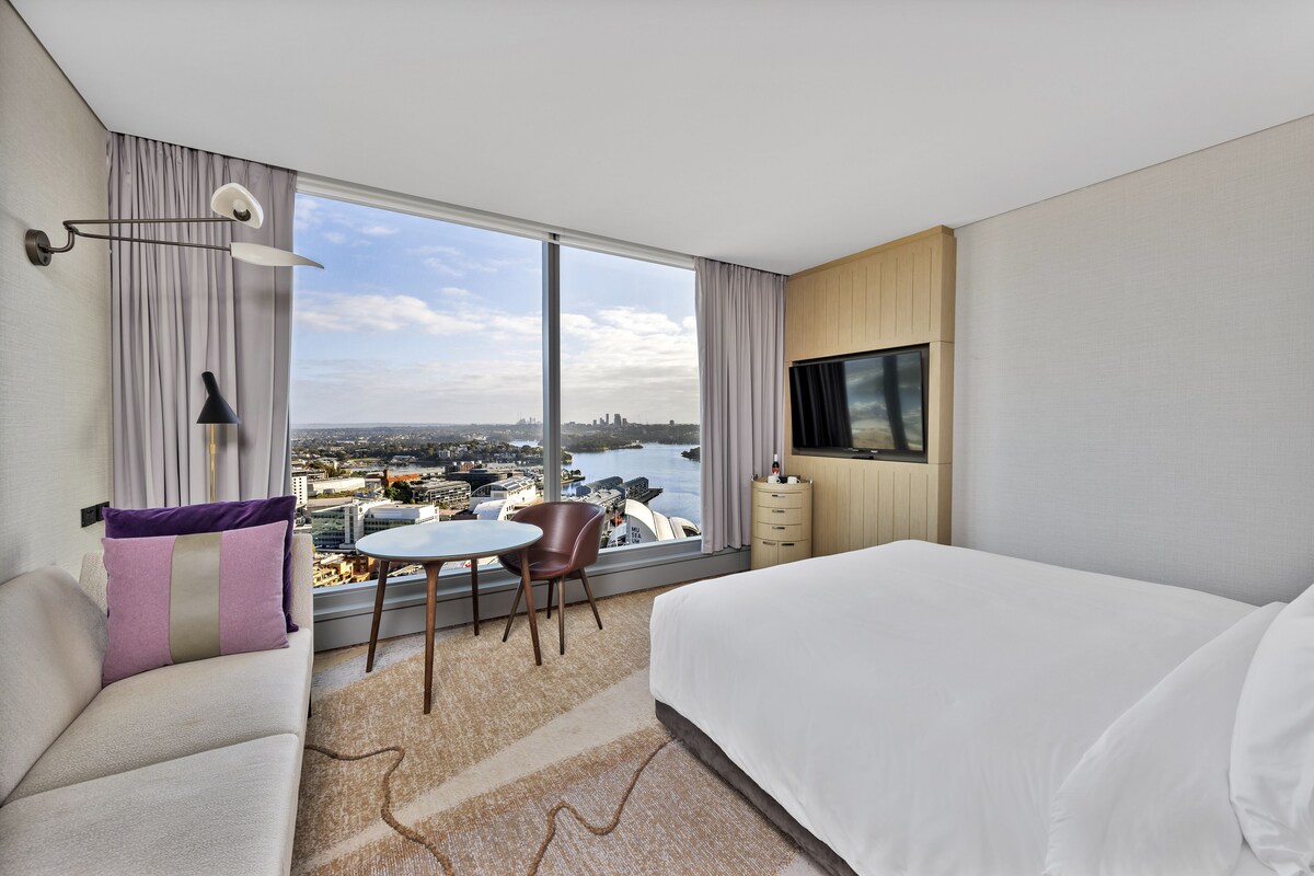Una habitación de hotel con un ventanal grande, una cama, un sofá, una mesita y una silla.