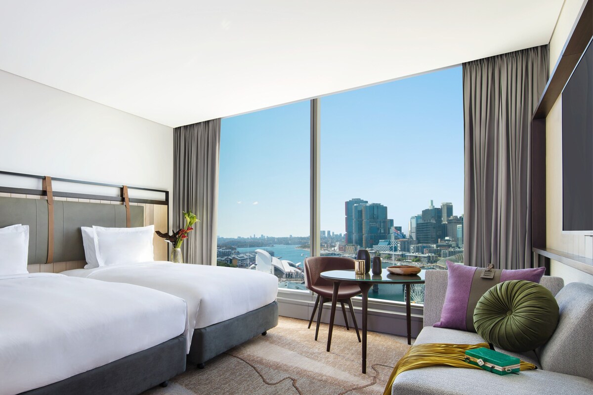 Habitación de hotel con dos camas, un sofá, una mesita y vistas a la silueta de la ciudad.