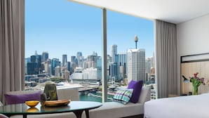 Habitación de hotel con un amplio ventanal que ofrece vistas a la ciudad, un sofá con un cojín de colores, una mesa de vidrio con un cuenco y un florero, y una cama con ropa de cama blanca.