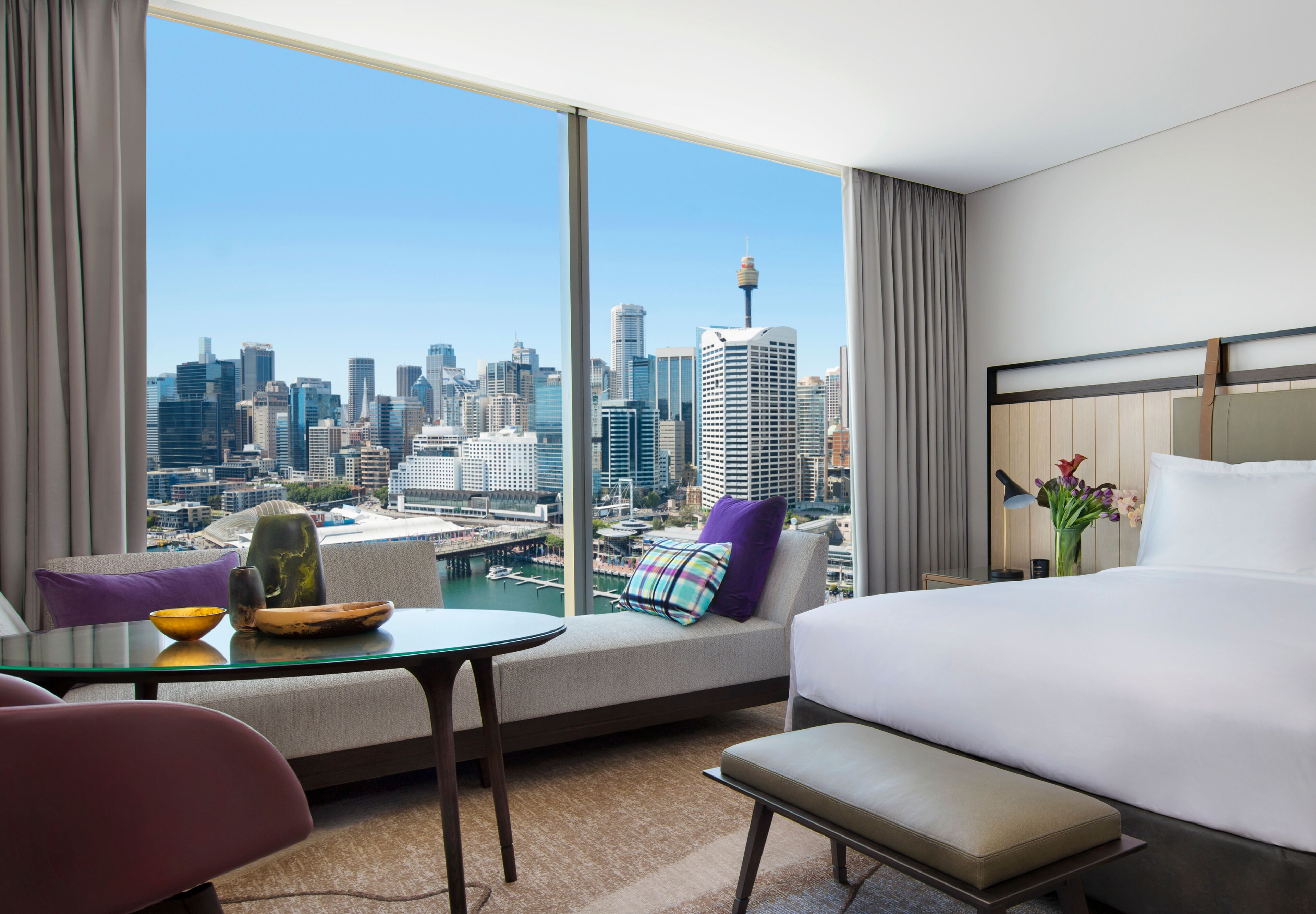 Habitación de hotel con un ventanal grande que ofrece vistas a la ciudad, una cama, un sofá, una mesita y un banco.