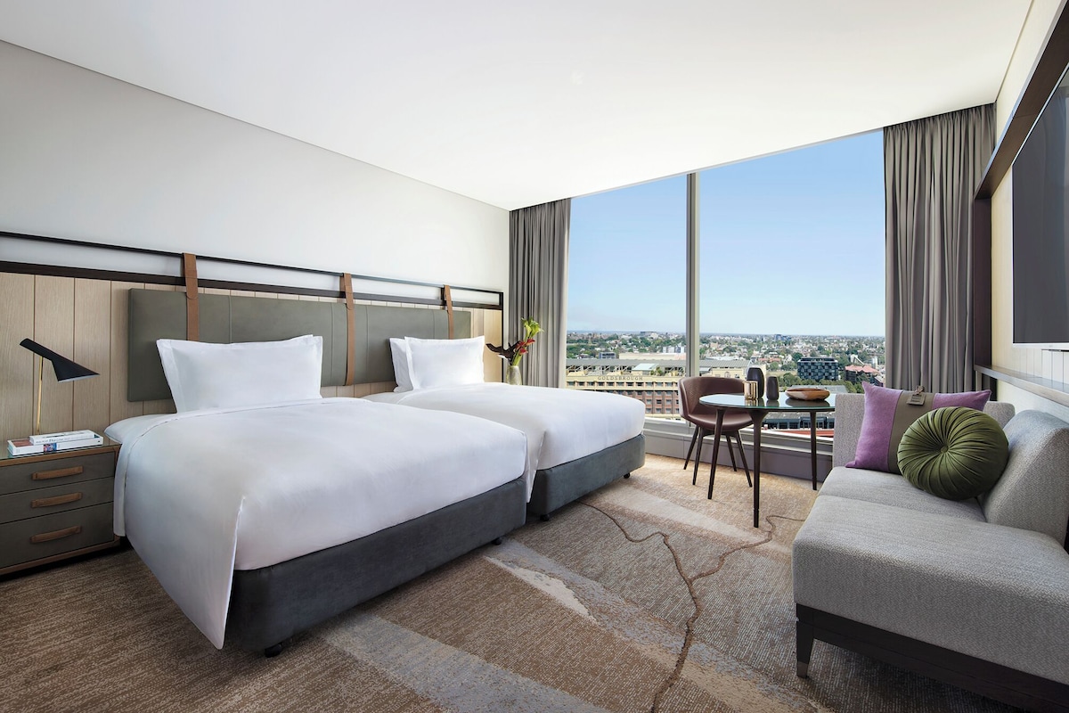 Habitación de hotel con dos camas, un sofá, una mesa de comedor y un ventanal con vistas a la ciudad.