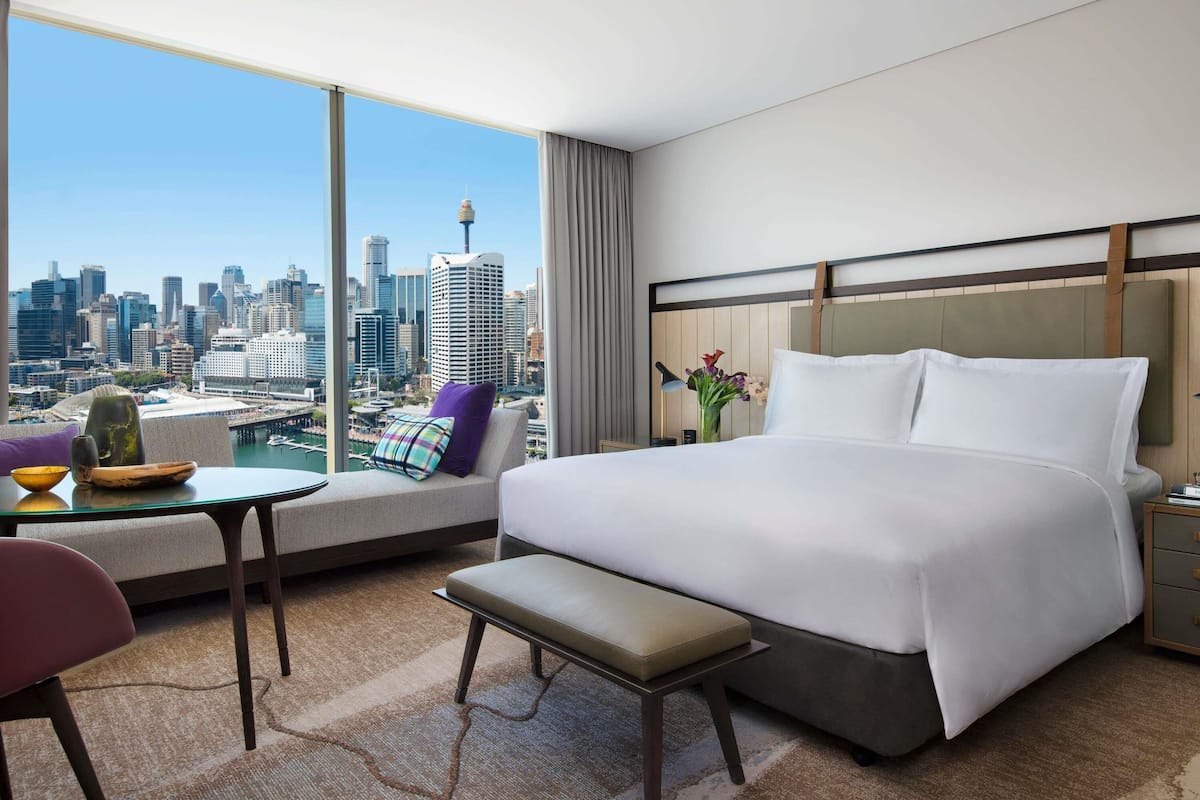 A modern hotel room with a large bed, a small table, a chair, and a view of a city skyline.