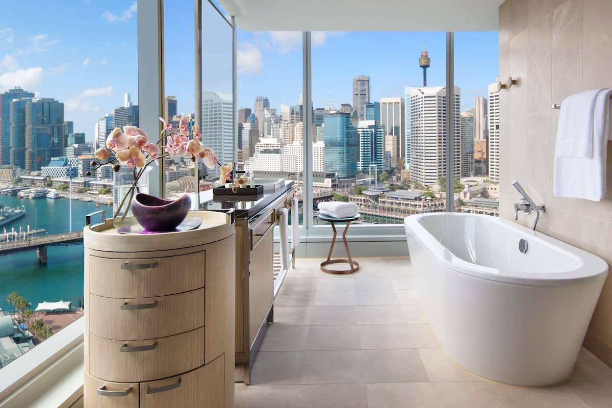 A modern bathroom with a bathtub, a wooden vanity, and a view of a city skyline and harbor.