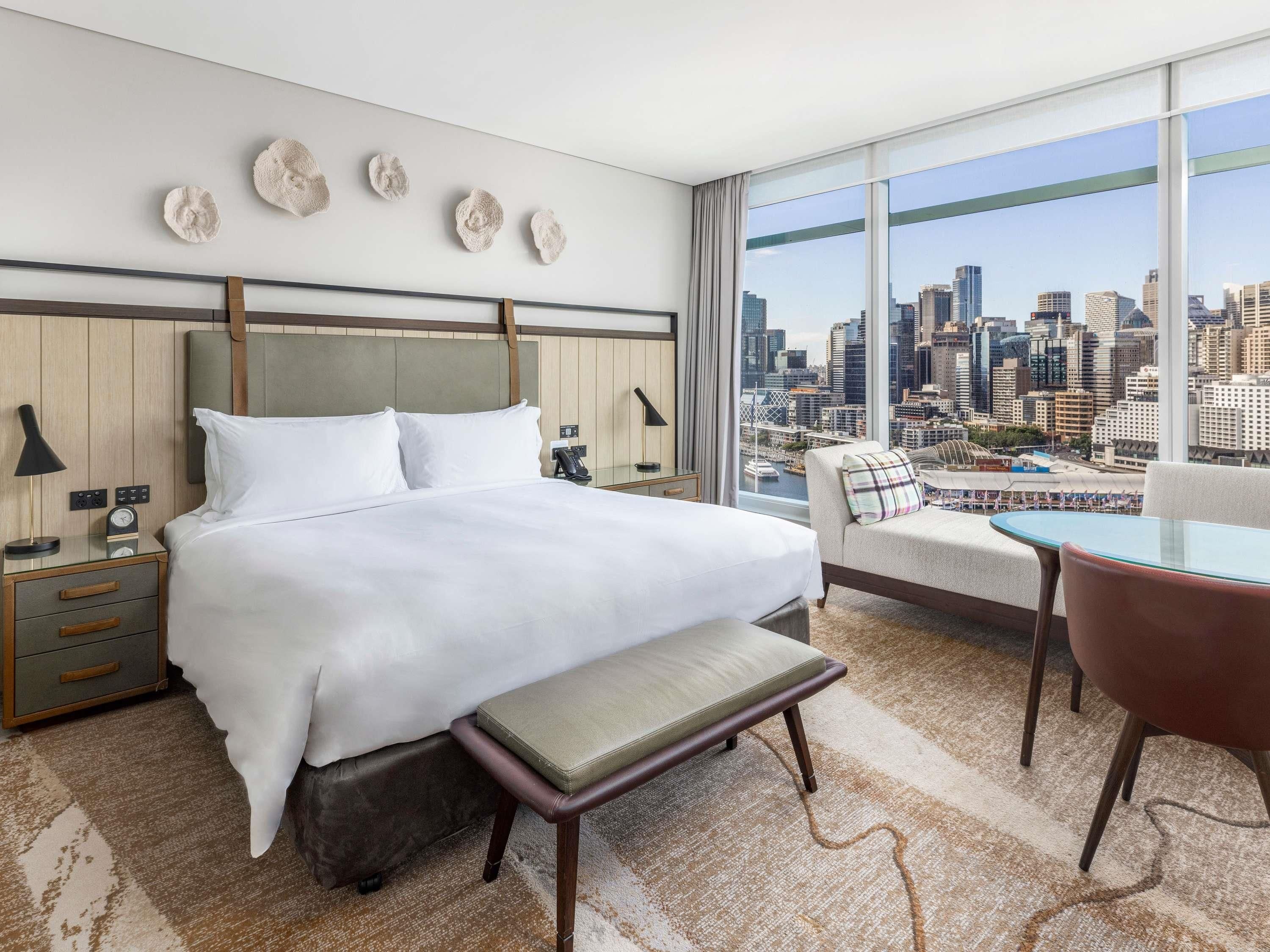 A hotel room with a large bed, a bench, a chair, a desk, and a city view.