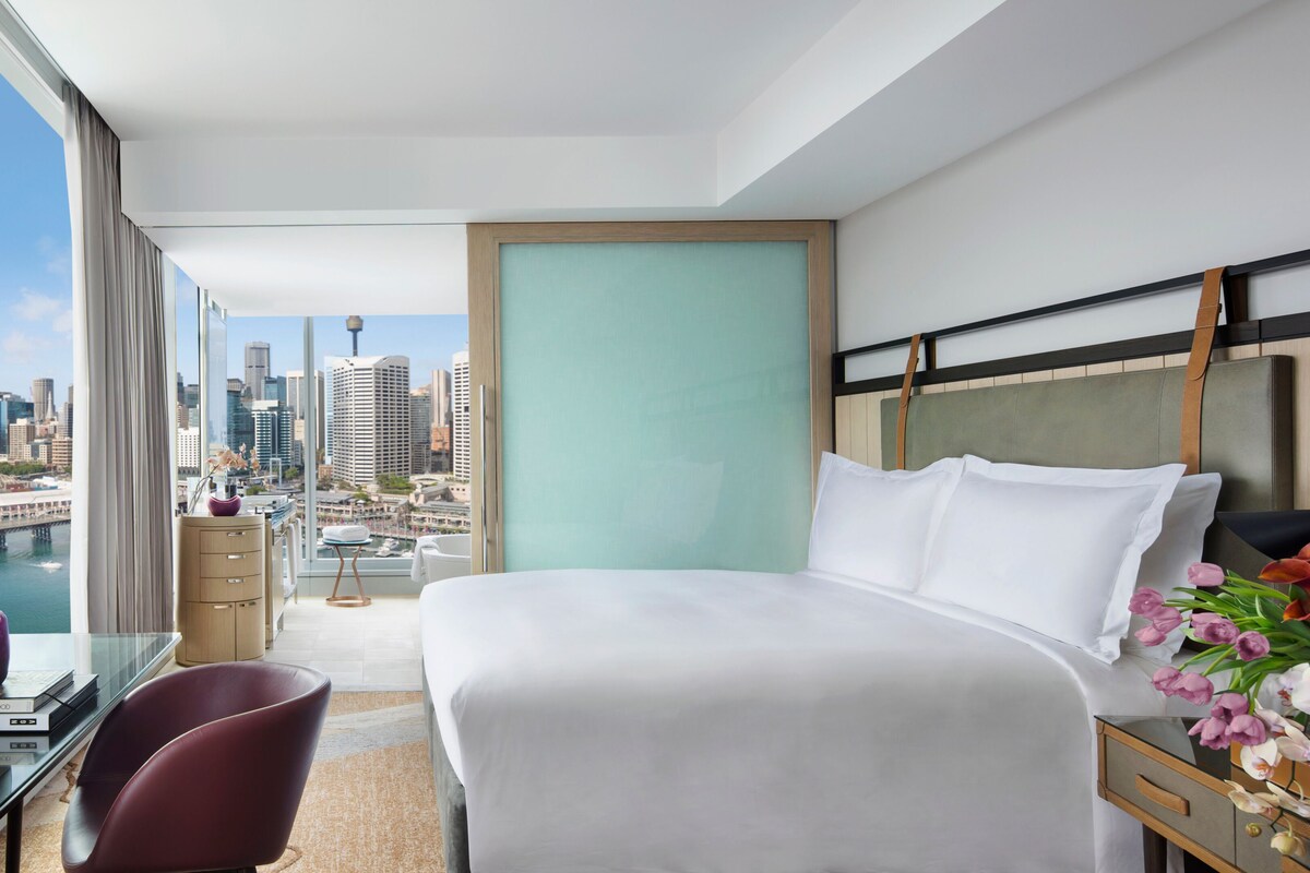 Una habitación de hotel con una cama grande, un escritorio con silla, vista a la ciudad y un horizonte urbano visible a través de la ventana.