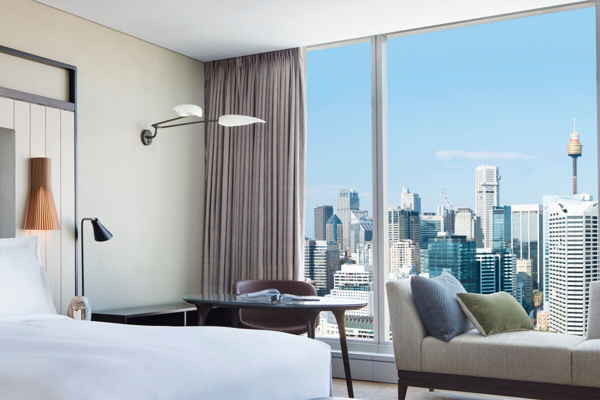 Habitación de hotel con cama, escritorio, silla, sofá y una ventana grande que ofrece vistas al horizonte de la ciudad.