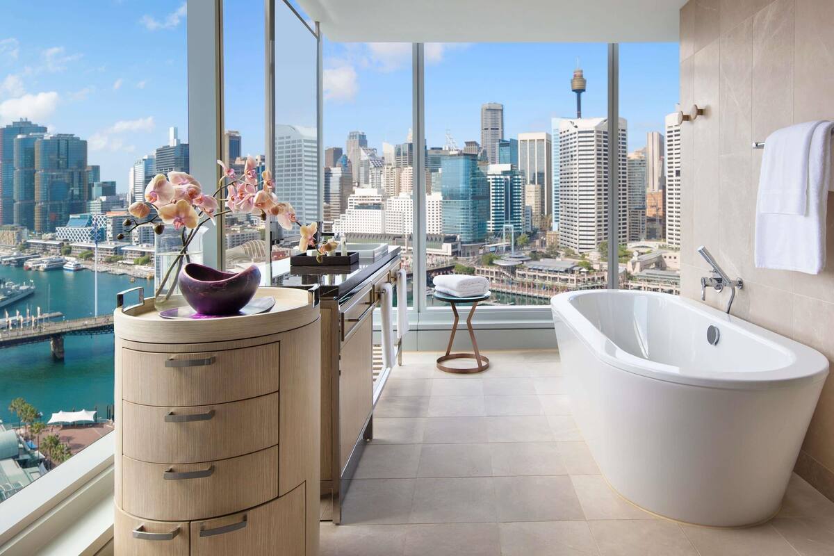 A modern bathroom with a bathtub, a wooden vanity, and a view of a city skyline and harbor.