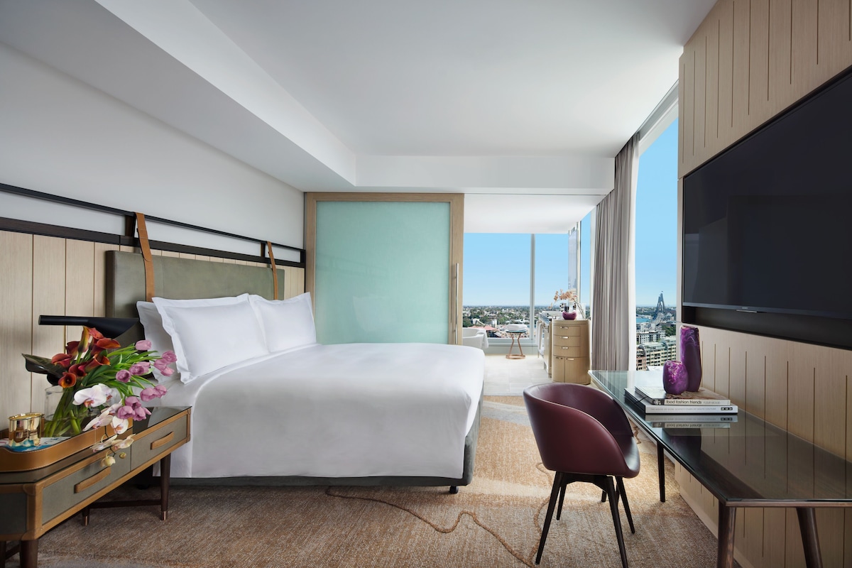 Una habitación de hotel moderna con una cama grande, un escritorio con silla, televisión y vista a la ciudad.