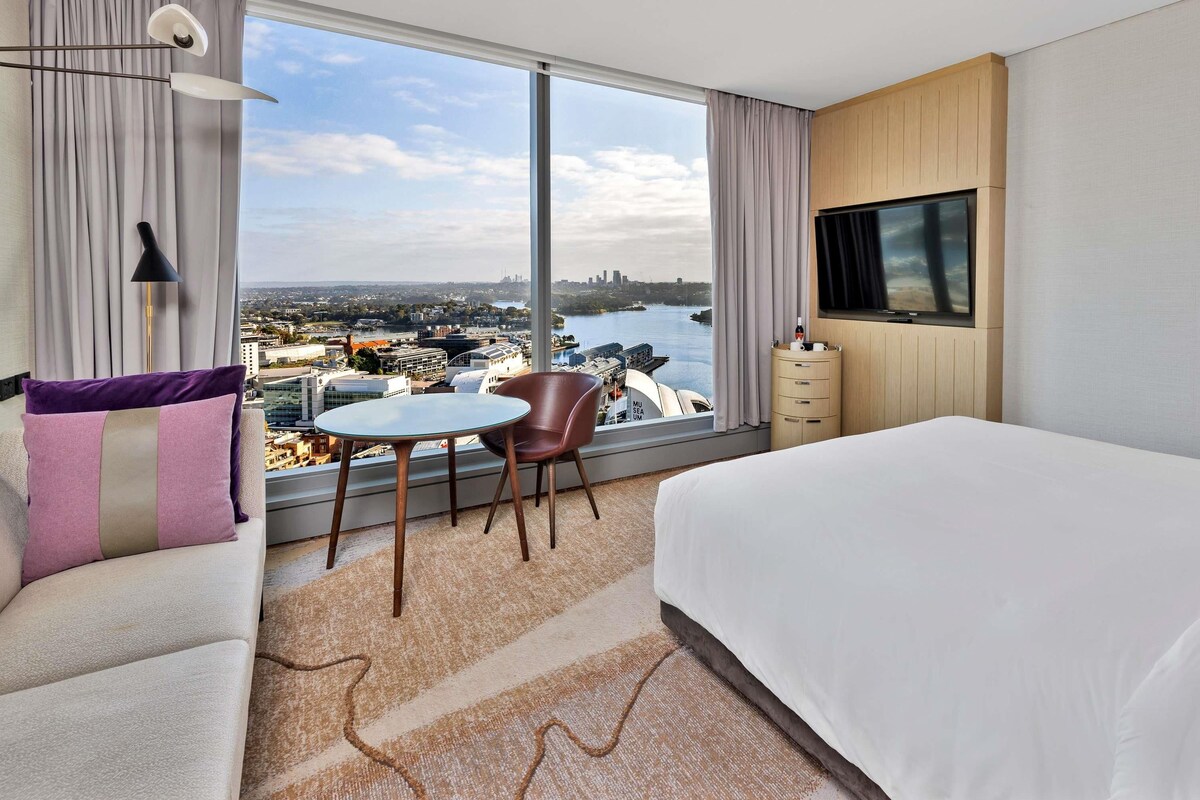 Habitación de hotel con una cama grande, un televisor en la pared, una mesa de comedor con sillas y vistas a un paisaje urbano a través de grandes ventanales.