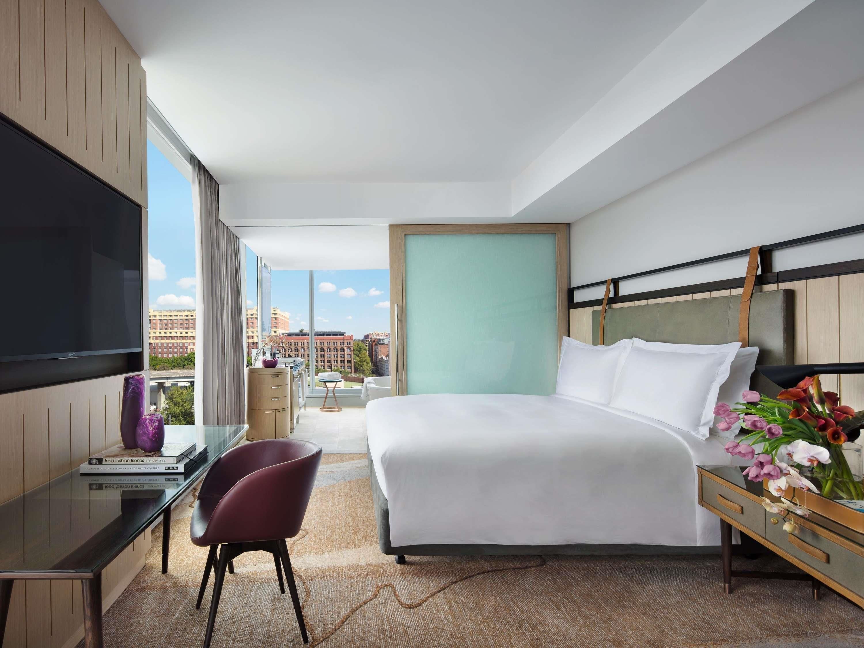 A modern hotel room with a large bed, a desk with a chair, a TV mounted on the wall, and a view of the cityscape.
