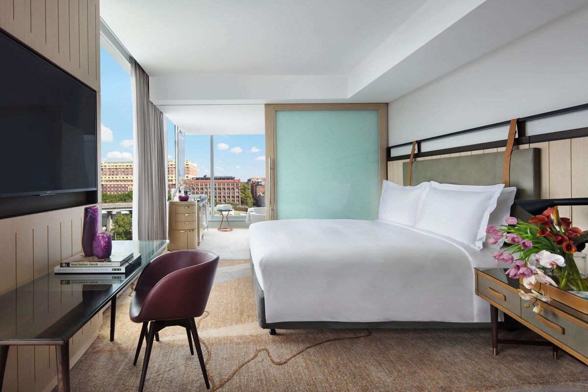 Habitación de hotel moderna con una cama grande, un escritorio con una silla, un televisor montado en la pared y vistas a la ciudad.