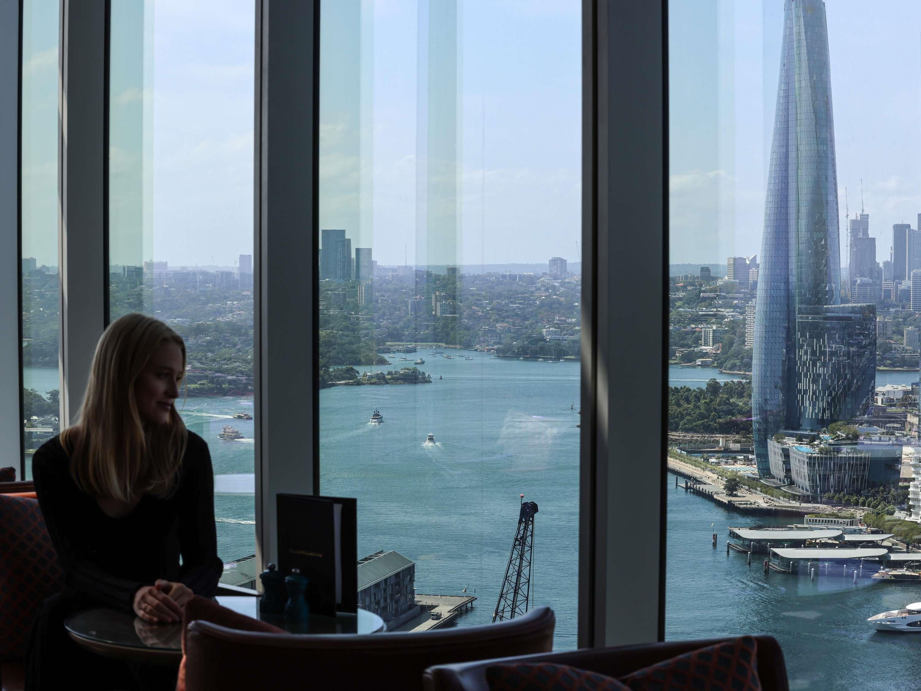 A person seated by a large window overlooking a cityscape with a river, a tall building, and a bridge.