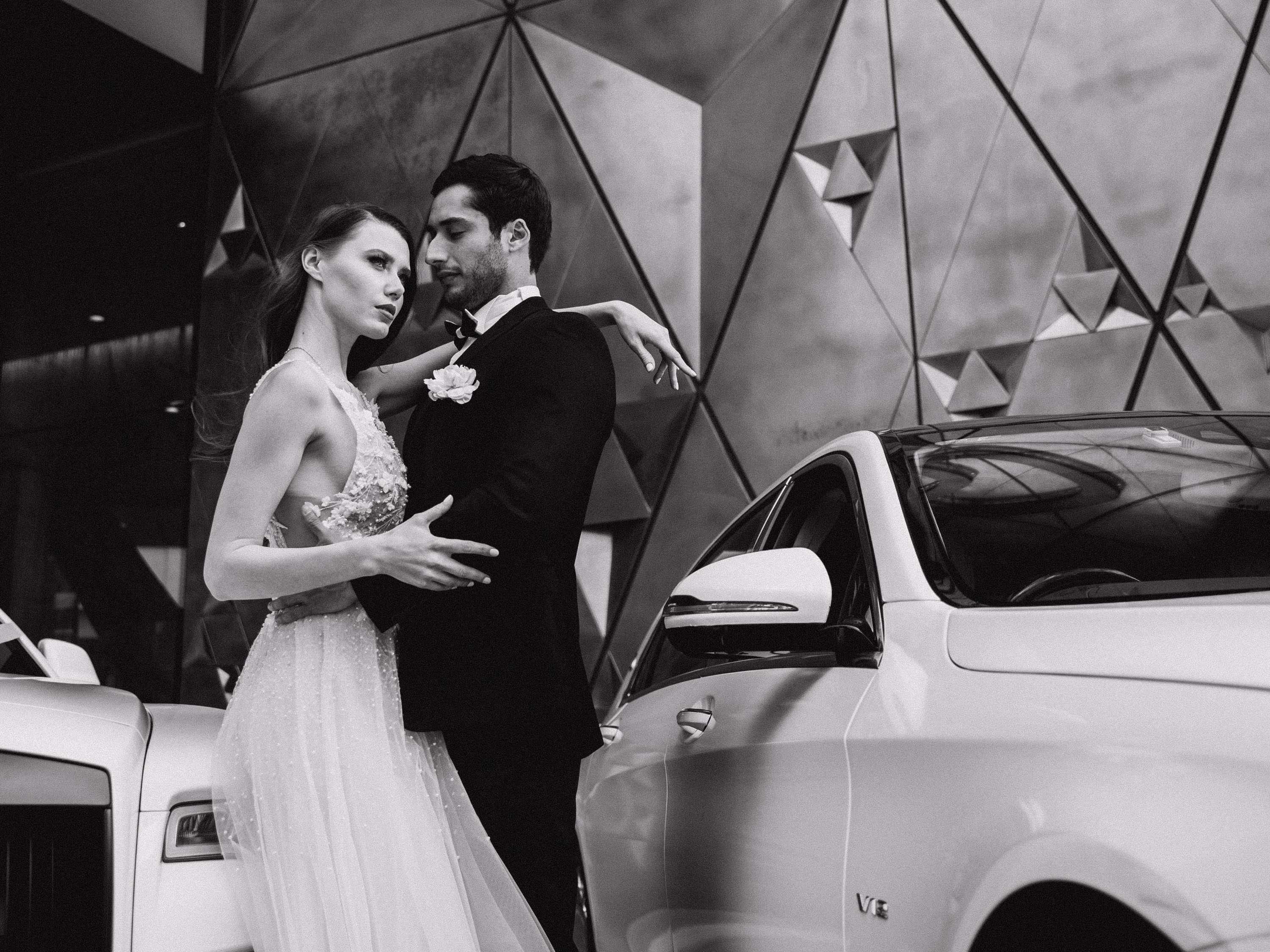 Una pareja posa junto a un coche con un fondo geométrico.