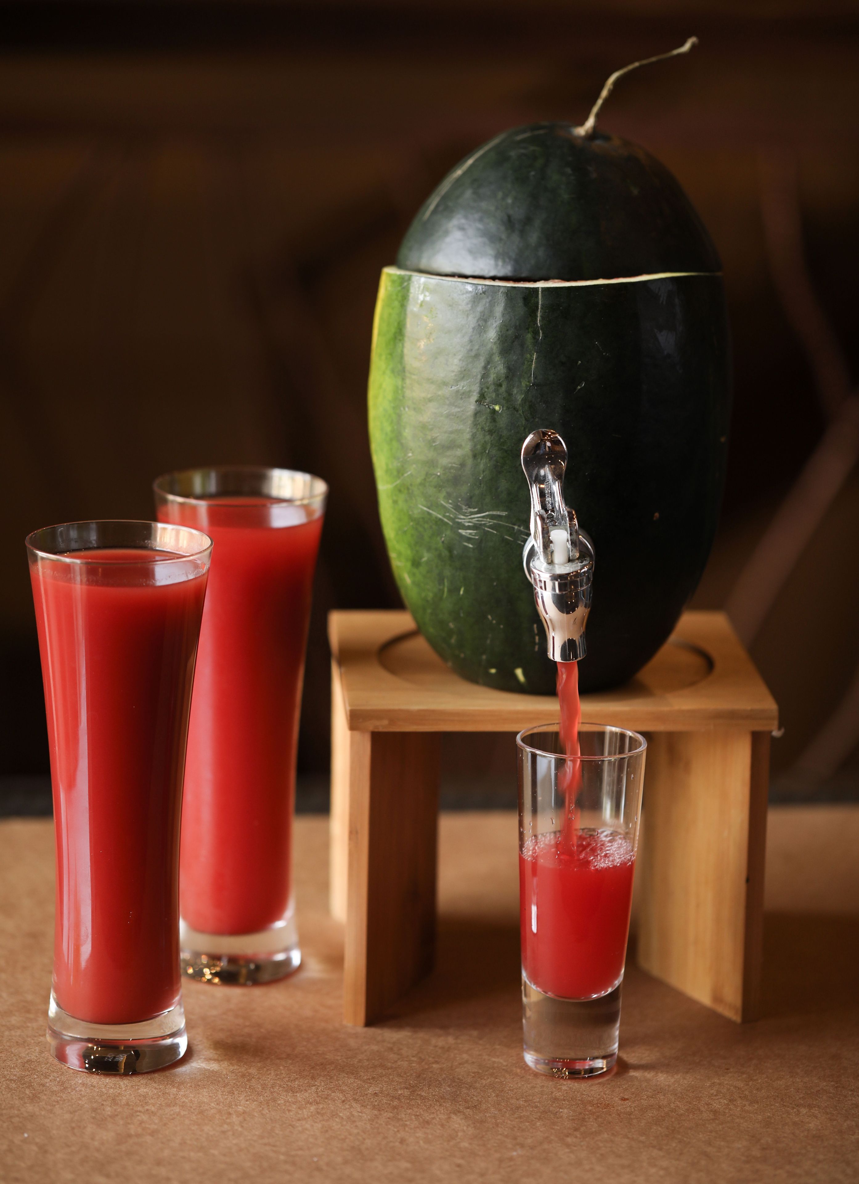 Una mesa de madera con un grifo metálico vertiendo un líquido rojo en un vaso, junto con otros dos vasos llenos del mismo líquido.